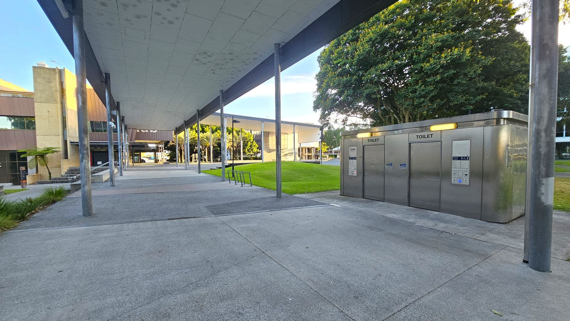 A modern outdoor area with a wide concrete pathway. On the right, there are stainless steel public toilets with illuminated signs. To the left, a building with large windows and a beige facade is visible. The ceiling above is flat and supported by metal poles. In the background, there is a grassy area with a large tree and another building with a similar architectural style. The area is well-lit and open, with some small plants along the pathway.