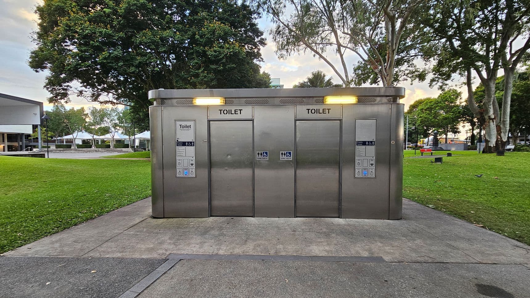 A modern public toilet facility with three stainless steel doors, each labelled "TOILET". The structure is set on a concrete path surrounded by a grassy area. Trees are visible in the background, providing a natural setting. The facility has illuminated signs above each door, and there are control panels beside each entrance. The area appears clean and well-maintained, with additional park features like benches and pathways visible in the distance.