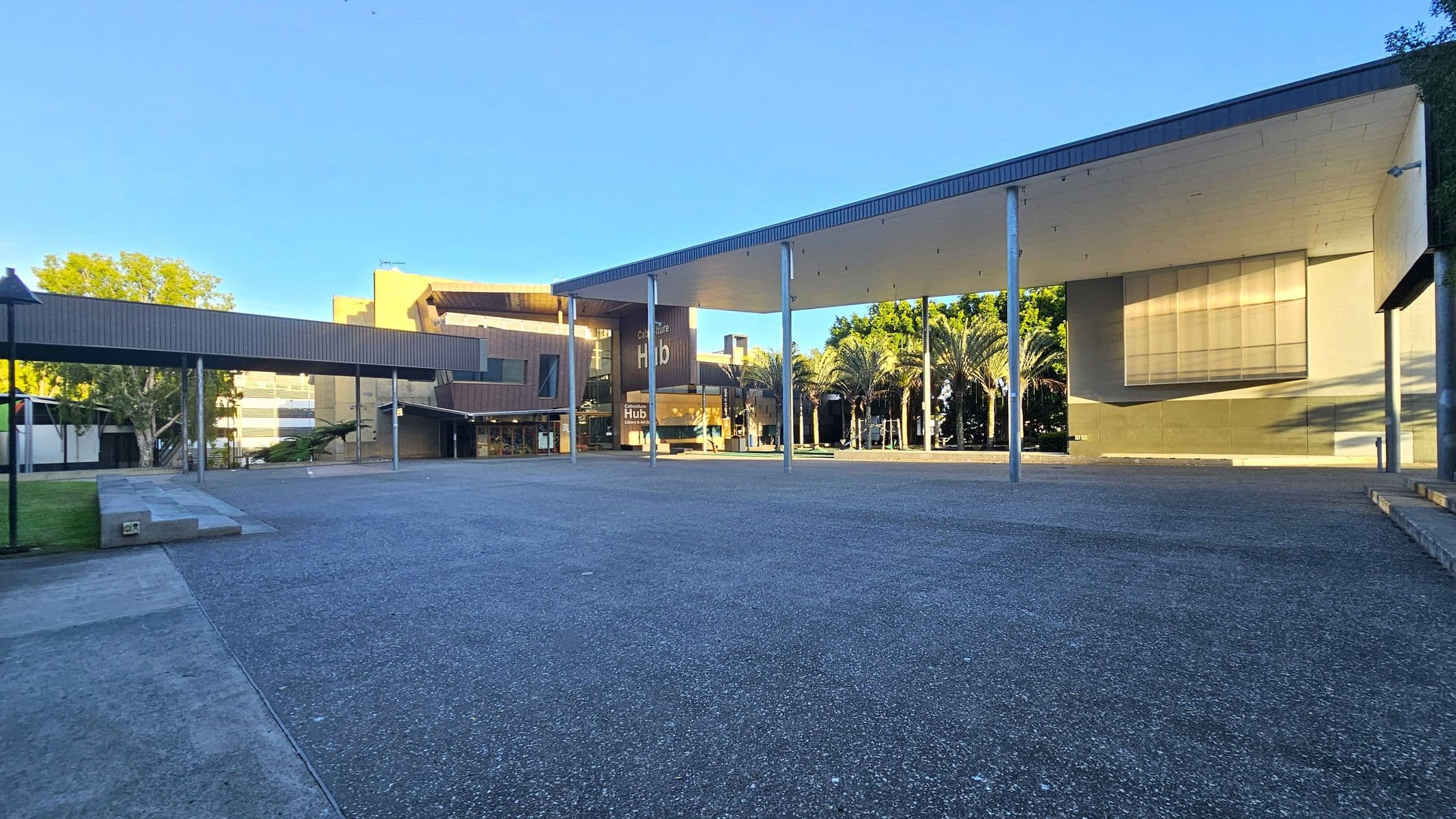 A spacious outdoor area with a modern architectural design. On the left, a long, covered walkway supported by metal poles leads to a building with a sign reading "Culture Hub." The building has a mix of glass and wooden panels. On the right, a large open space with a high ceiling supported by columns. Palm trees are visible in the background, adding greenery. The ground is paved with a mix of concrete and textured surfaces.