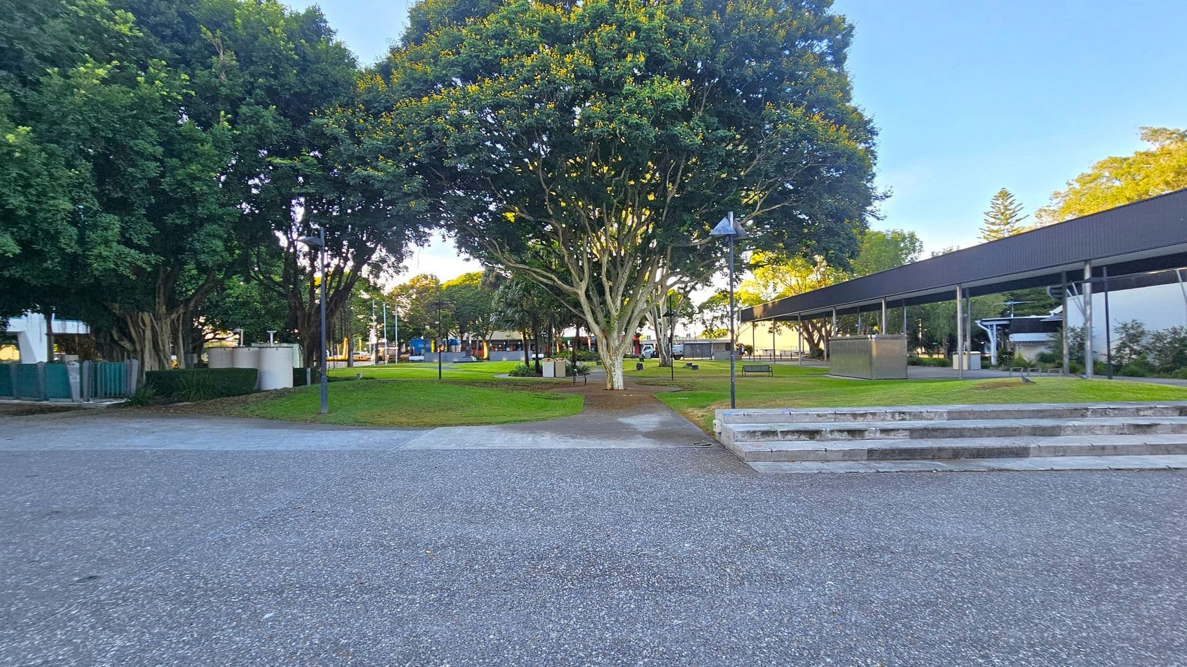A park scene with a large tree in the centre, surrounded by green grass and pathways. On the left, there are more trees and a small structure with a green fence. On the right, a covered walkway with a metal roof extends into the distance. In the foreground, there is a paved area with steps leading up to the grassy section. Benches and lampposts are scattered throughout the park.