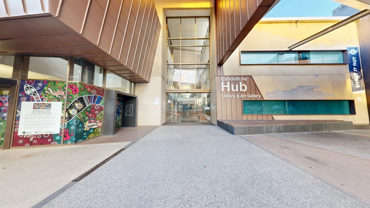 The entrance to the Caboolture Hub Library & Art Gallery features a modern architectural design. On the left, there is a colourful mural with abstract patterns and a white information board. The centre has large glass doors and windows, framed by a metallic structure. On the right, the building's name is displayed on a brown panel, with narrow horizontal windows below. The ground is paved with light grey concrete, and the ceiling has a metallic finish.