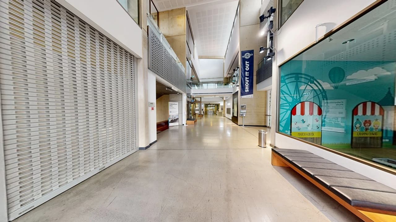 A spacious indoor corridor with a polished floor. On the left, a large metal shutter is closed. On the right, a glass display with colourful graphics and a bench below it. The ceiling is high with visible lighting fixtures. In the centre, a banner hangs with the words "SHOUT IT OUT". The corridor extends into the distance with various signs and a few bins along the walls. The upper level features glass railings.