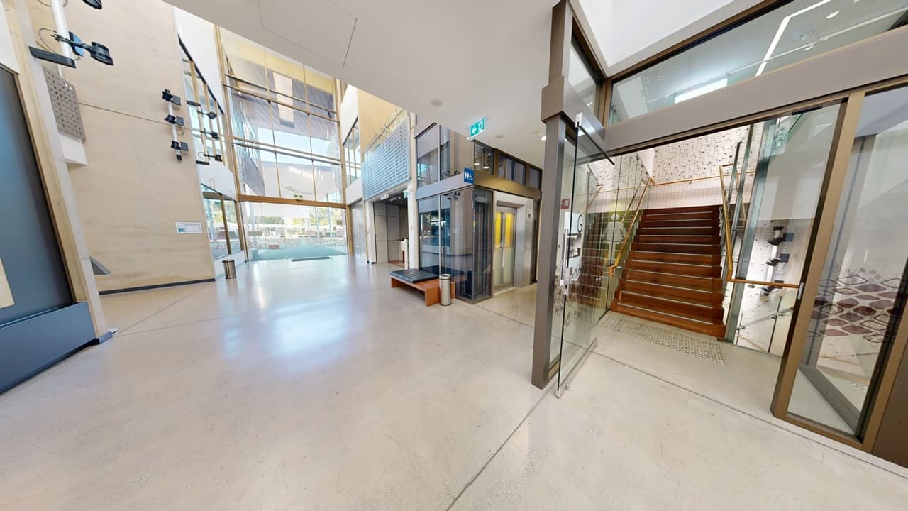 Spacious modern interior with a light-coloured floor. On the left, a wall with mounted objects and large windows allowing natural light. In the centre, a glass lift with a wooden bench nearby. On the right, a staircase with wooden steps and glass railings. The ceiling is high with a clean, minimalist design.