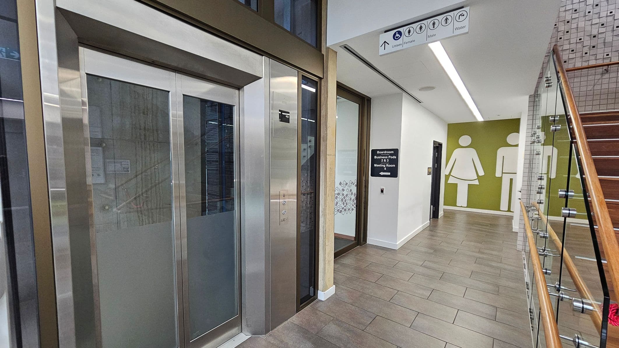 On the left, there is a modern lift with metallic doors. To the right, a corridor leads to restrooms, indicated by large male and female symbols on a green wall. Above, a sign directs to unisex, female, male, and accessible toilets. A staircase with glass panels and a wooden handrail is on the far right. The floor is tiled in a mix of brown and grey, and the ceiling features recessed lighting.