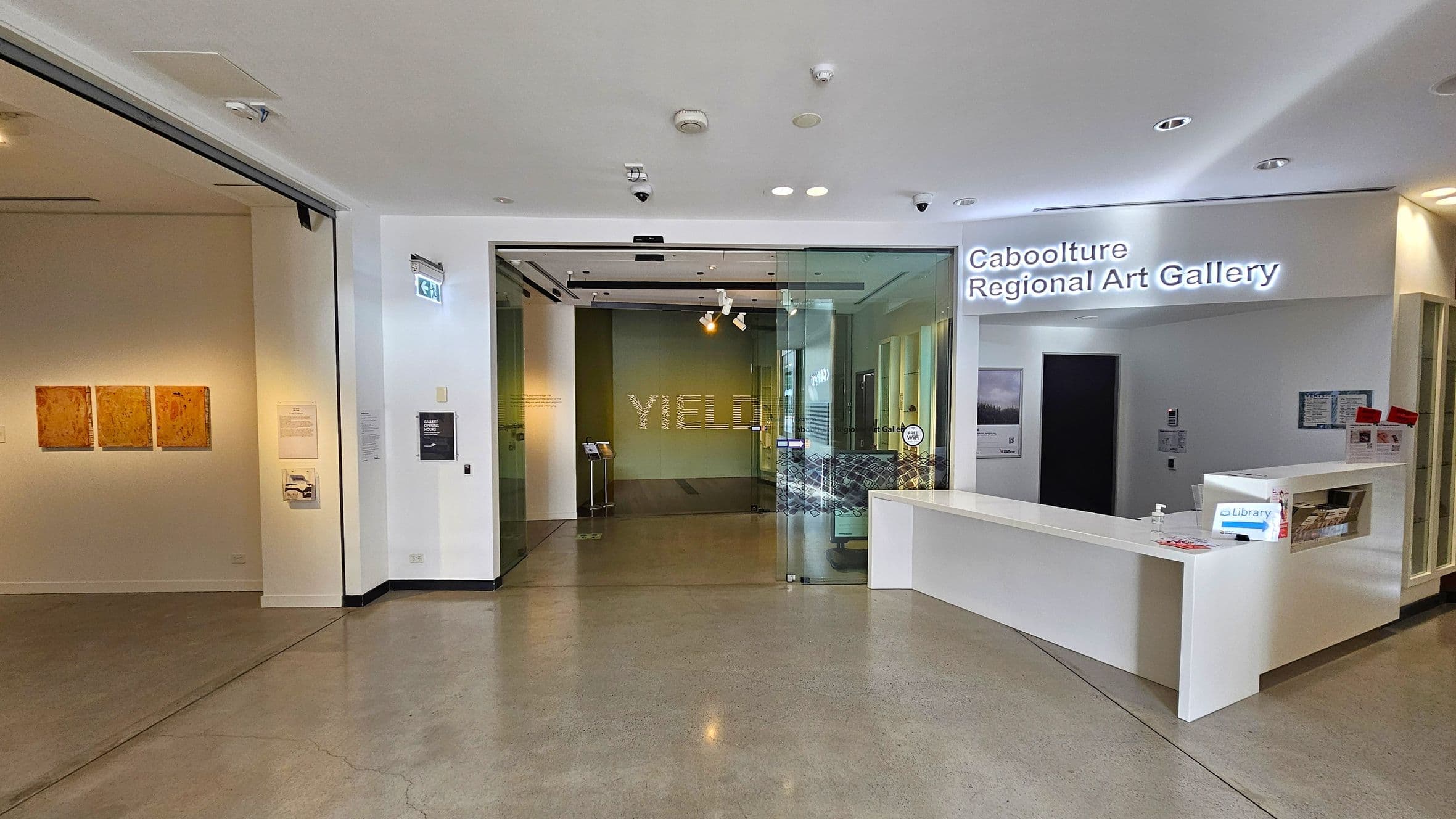 Entrance to the Caboolture Regional Art Gallery. On the left, a wall displays three square artworks. In the centre, a glass door leads into the gallery, with the word "YIELD" visible inside. On the right, a white reception desk with brochures and a hand sanitiser bottle. Above, a sign reads "Caboolture Regional Art Gallery". The floor is polished concrete, and the ceiling is white with recessed lighting.
