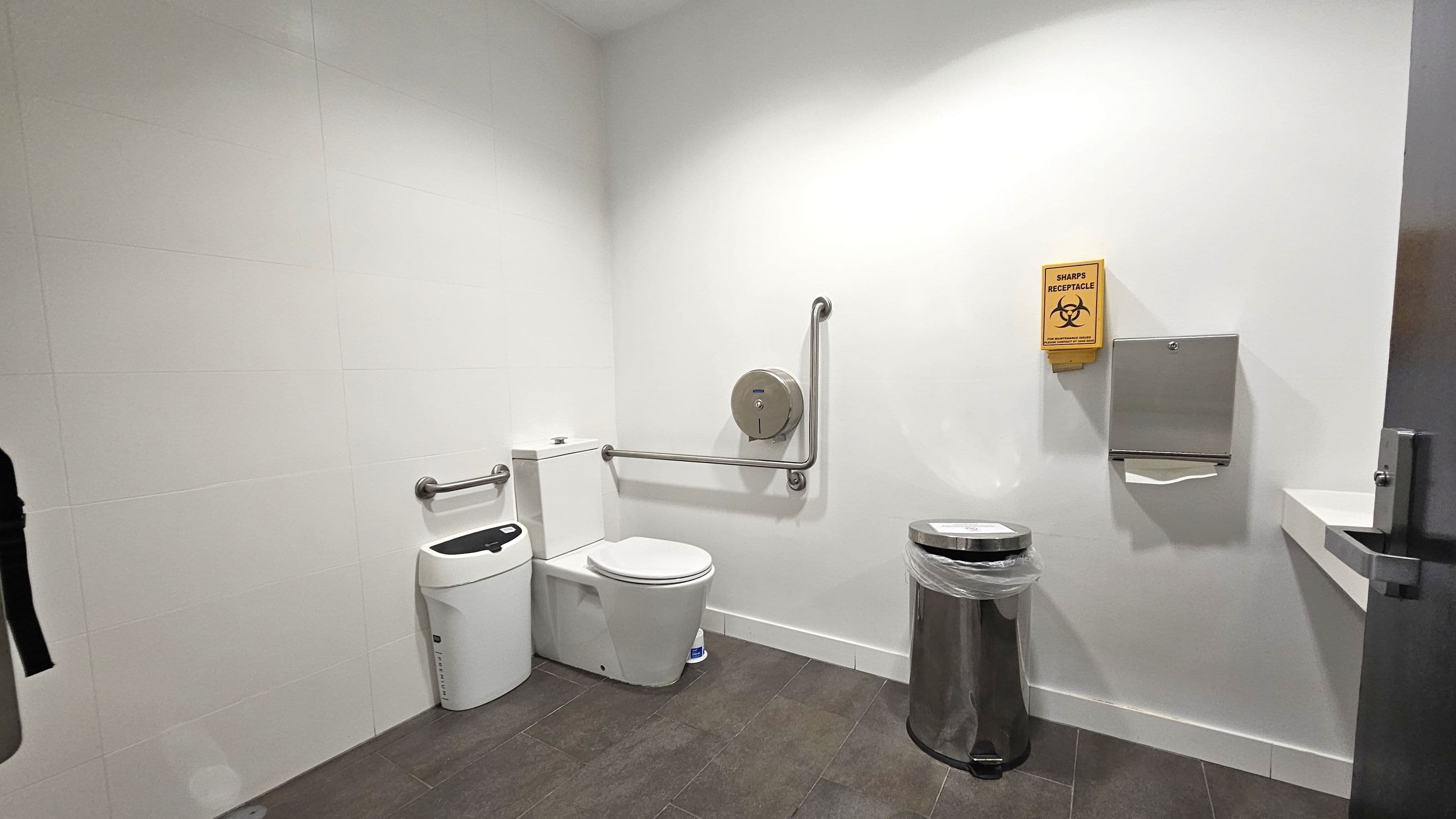 A clean, accessible bathroom with white walls and a dark tiled floor. On the left, there is a toilet with a support rail and a sanitary bin beside it. In the centre, a toilet paper dispenser is mounted on the wall. On the right, a metal bin with a liner, a sharps receptacle, and a paper towel dispenser are visible. A small sink is partially visible on the far right, next to the door.