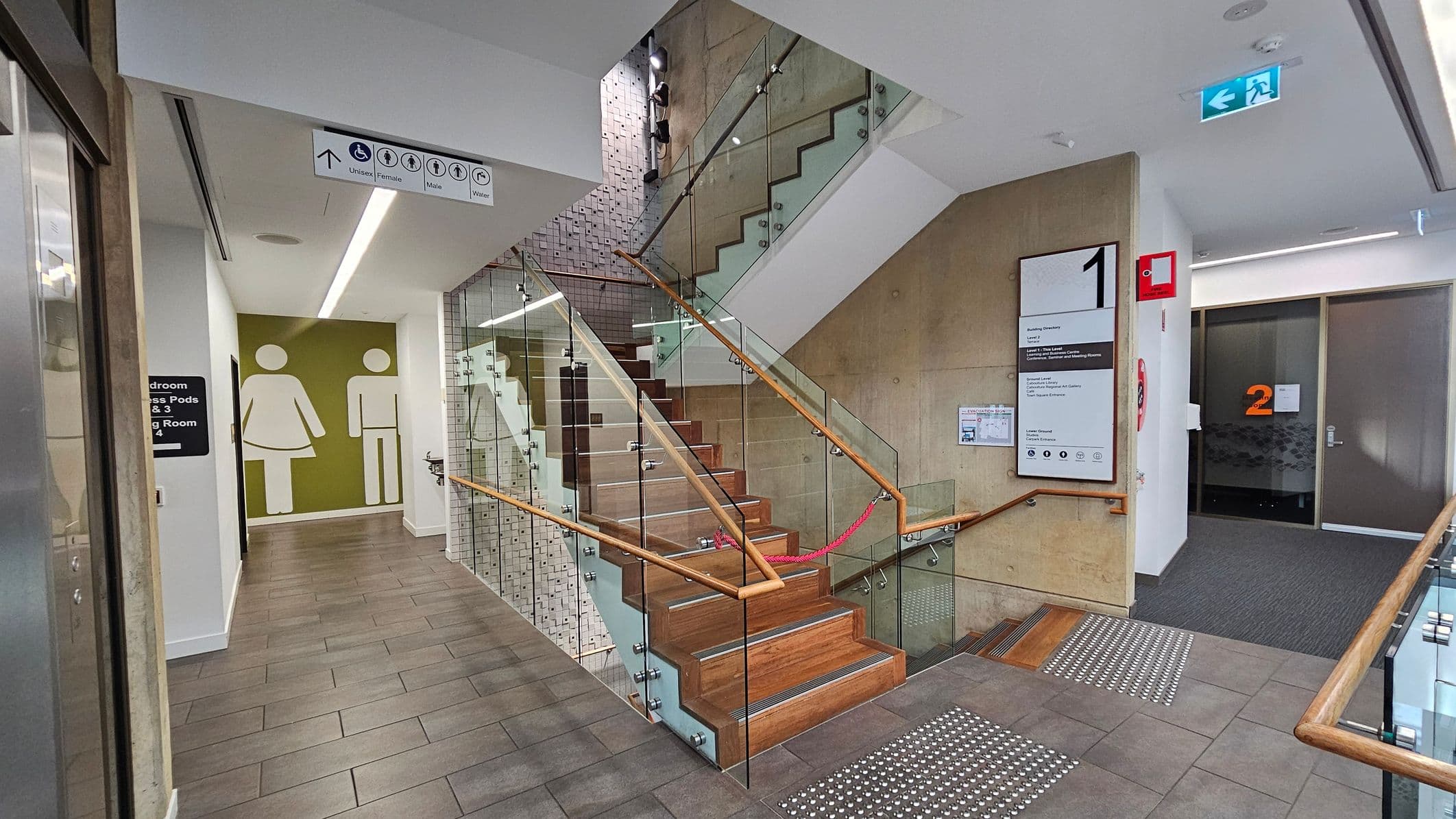 A modern indoor space with a staircase in the centre featuring wooden steps and glass railings. To the left, a hallway leads to restrooms, indicated by large male and female symbols on a green wall. Above, a sign shows restroom icons. On the right, a wall-mounted directory and a fire extinguisher are visible. The floor is tiled, and the ceiling has recessed lighting. An exit sign is mounted on the ceiling.