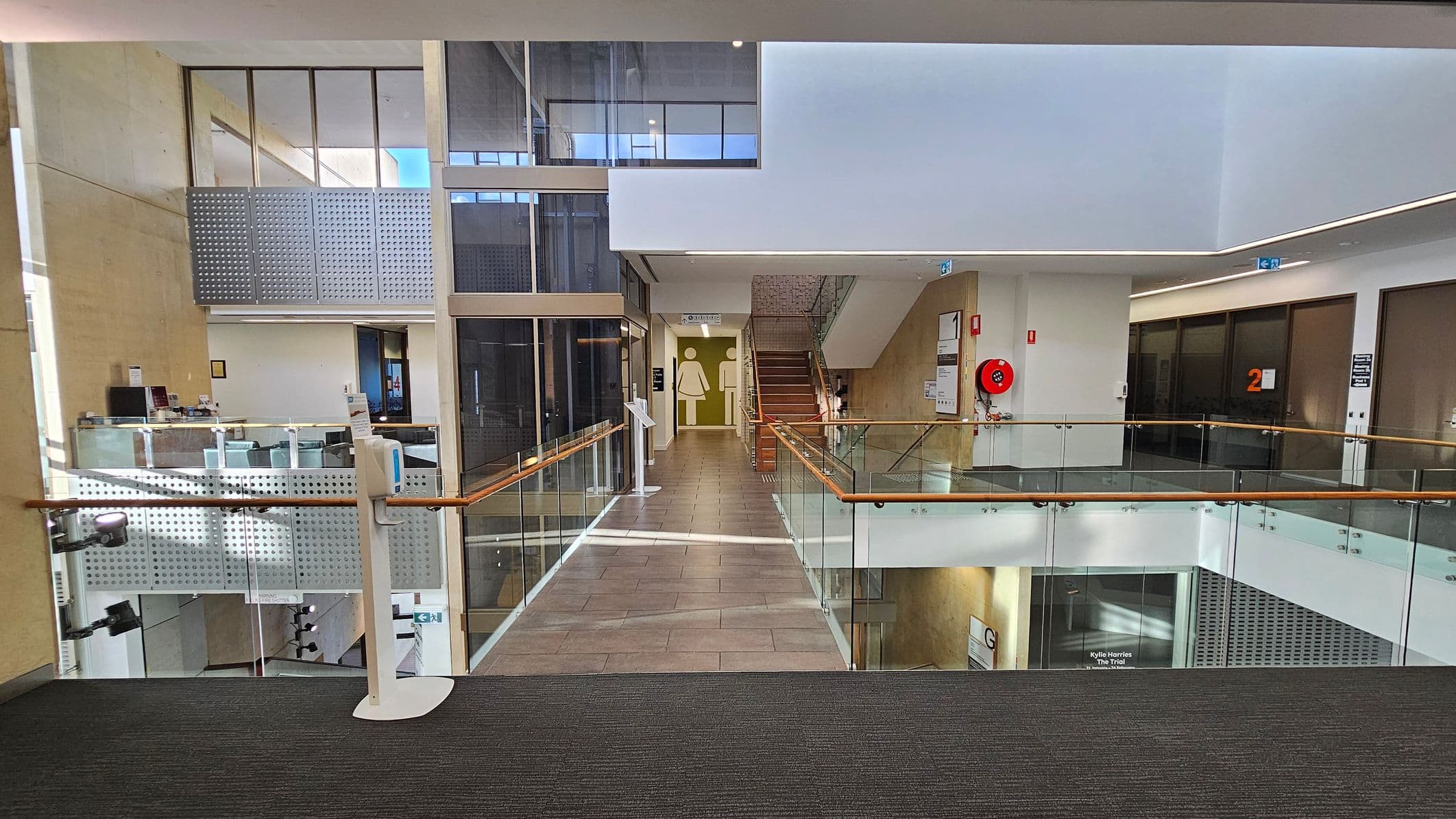 A modern interior with a carpeted floor in the foreground. A glass railing with wooden handrails lines the walkway. On the left, there are large windows and a reception area with a counter. In the centre, a corridor leads to a staircase, with restroom signs visible. On the right, there are doors marked with the number "2" and a red fire hose reel mounted on the wall. The ceiling is smooth with recessed lighting.