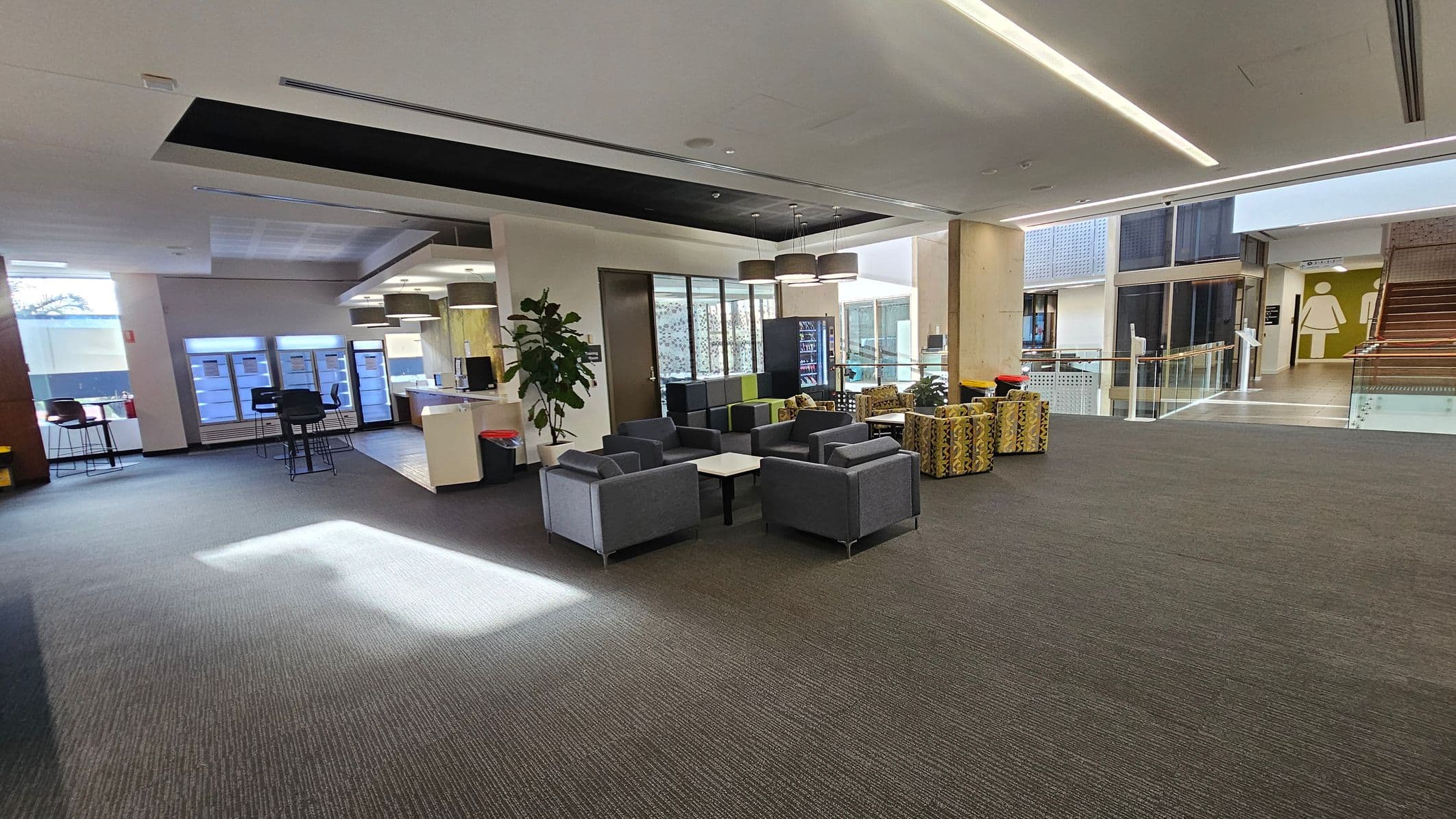 A spacious indoor area with a modern design. On the left, there are high tables and chairs near large windows. In the centre, a seating area with grey armchairs and a small table is surrounded by a few potted plants. To the right, a vending machine and patterned stools are visible. The background features a staircase leading to an upper level, with a large restroom sign on the wall. The floor is carpeted, and the ceiling has recessed lighting.