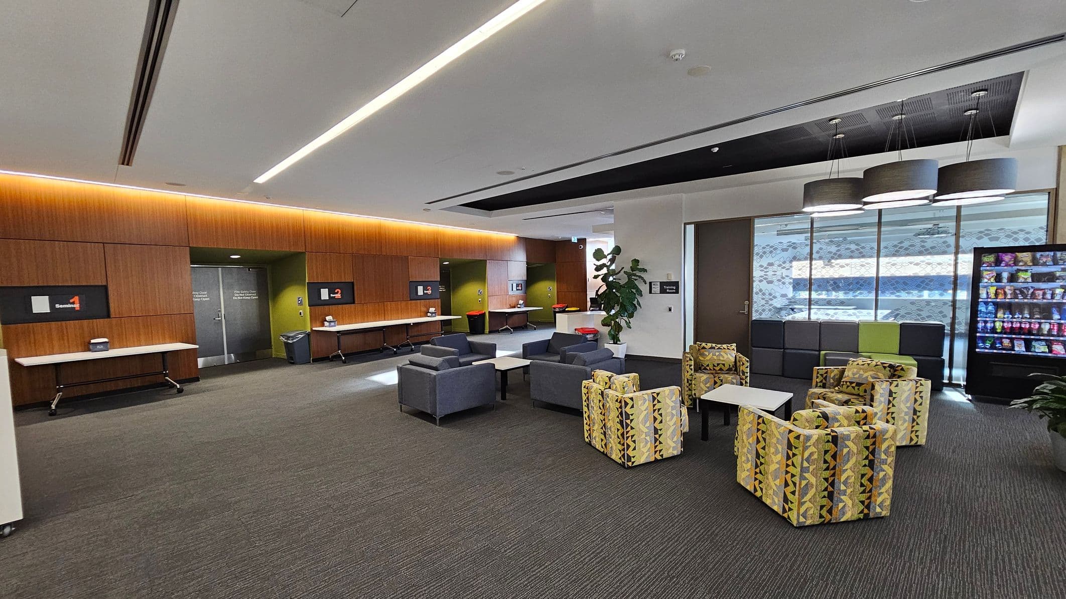 A modern waiting area with a grey carpeted floor. On the left, there are wooden panelled walls with digital screens and folding tables. In the centre, there are clusters of grey and patterned armchairs around small tables. On the right, a vending machine is next to a row of dark cushioned seats. Above, there are large circular ceiling lights. A potted plant is near the seating area, and large windows with patterned glass are in the background.