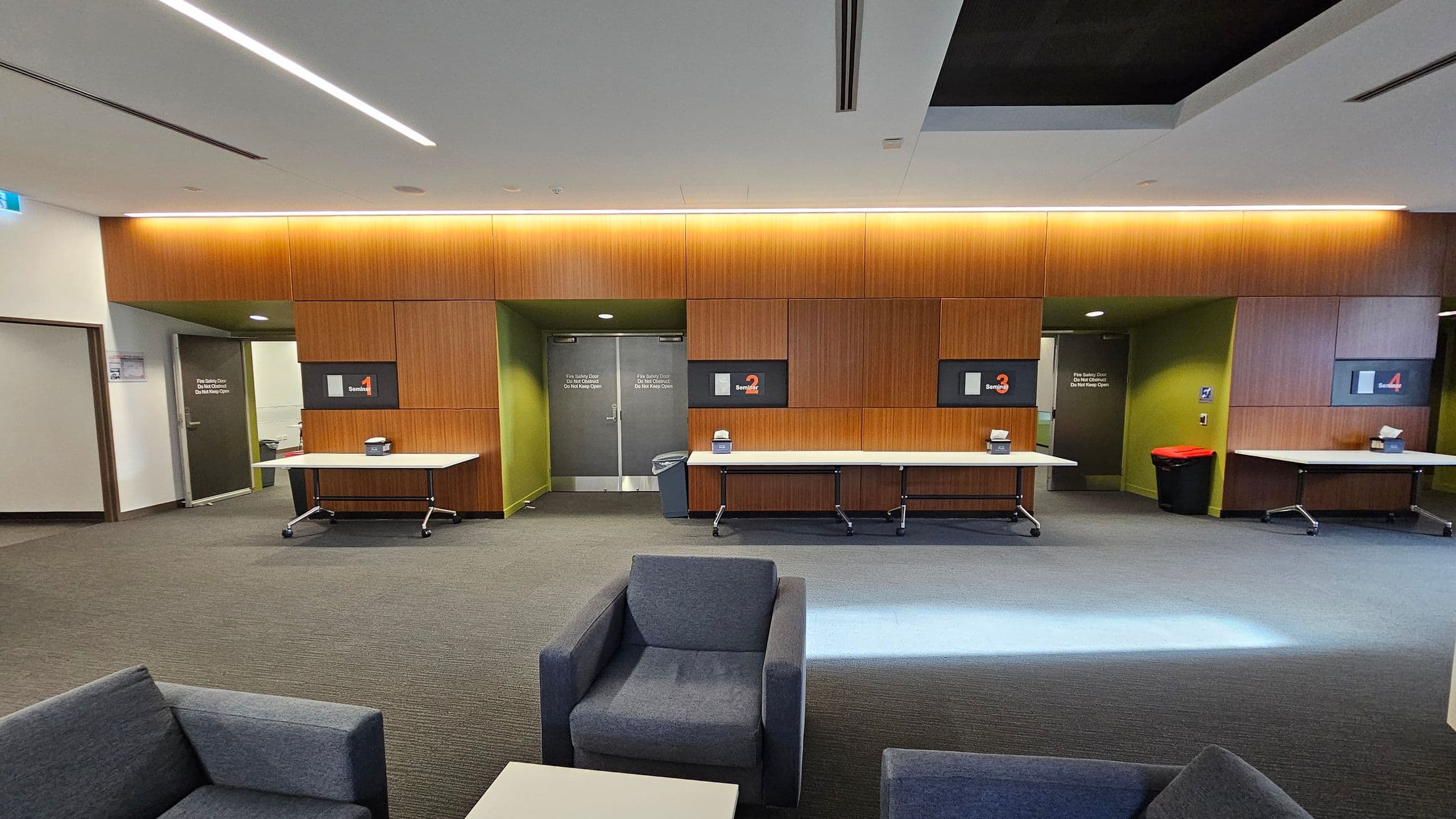 A modern interior with a row of wooden-panelled sections labelled "Section 1" to "Section 4" on the back wall. Each section has a white table with a box on top. The floor is carpeted in grey, and there are grey armchairs in the foreground. The ceiling features recessed lighting. Doors with signs are visible between the sections, and a red bin is near the right door.