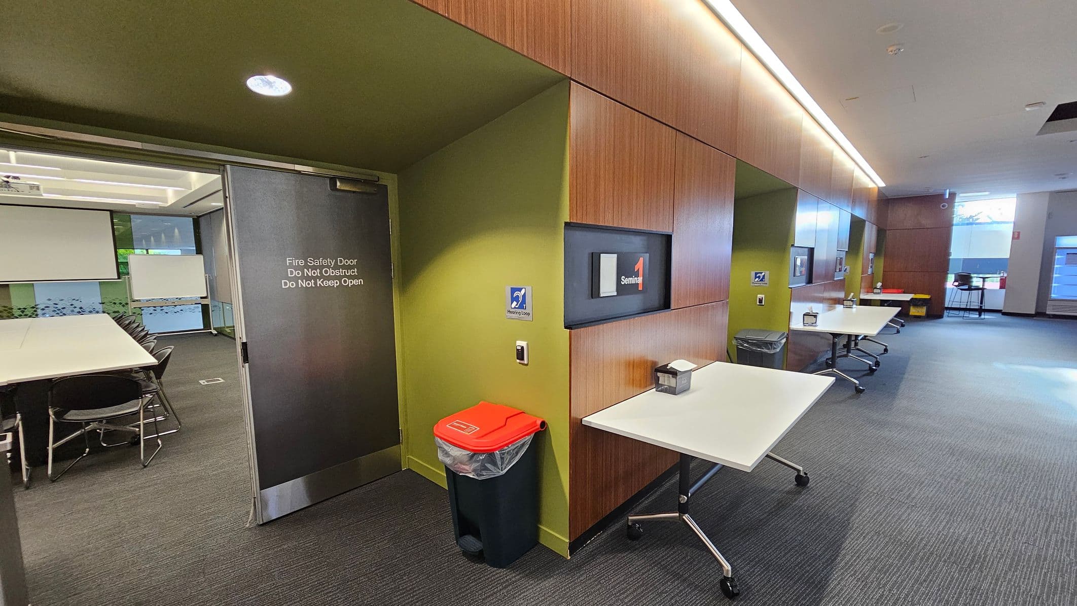 A modern interior space with a grey fire safety door on the left, leading to a room with tables and chairs. The wall is green with wood panelling, featuring a sign for "Seminar 1". A red-lidded bin and a small table with a tissue box are nearby. The right side has more tables and chairs, with large windows allowing natural light. The floor is carpeted, and the ceiling has recessed lighting.