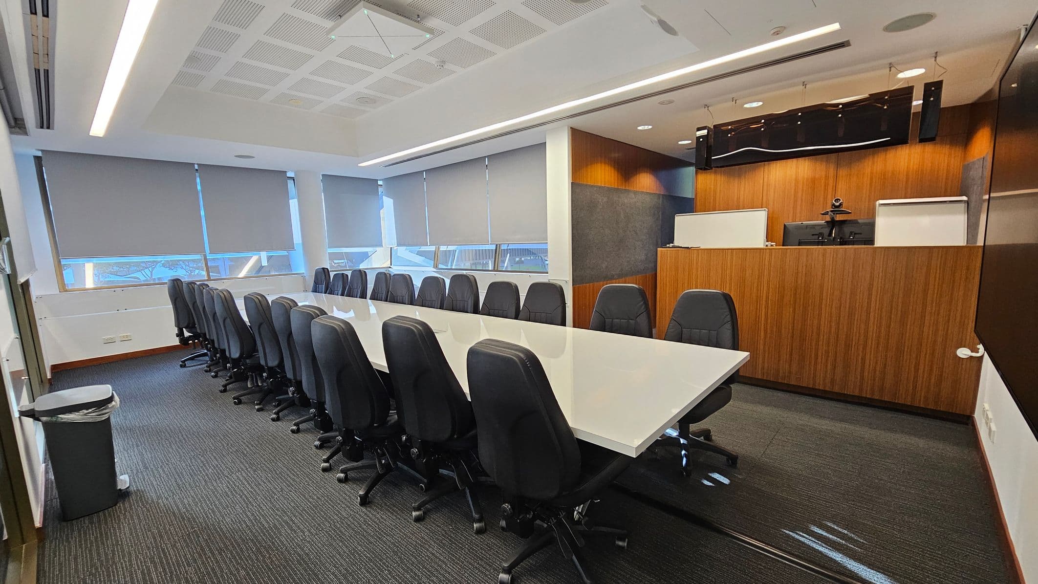 A modern conference room with a long white table surrounded by black office chairs. On the left, large windows with grey blinds allow natural light. The right side features a wooden panelled wall with a mounted screen and video conferencing equipment. The floor is carpeted in dark grey, and the ceiling has recessed lighting and acoustic panels. A waste bin is visible in the foreground on the left.