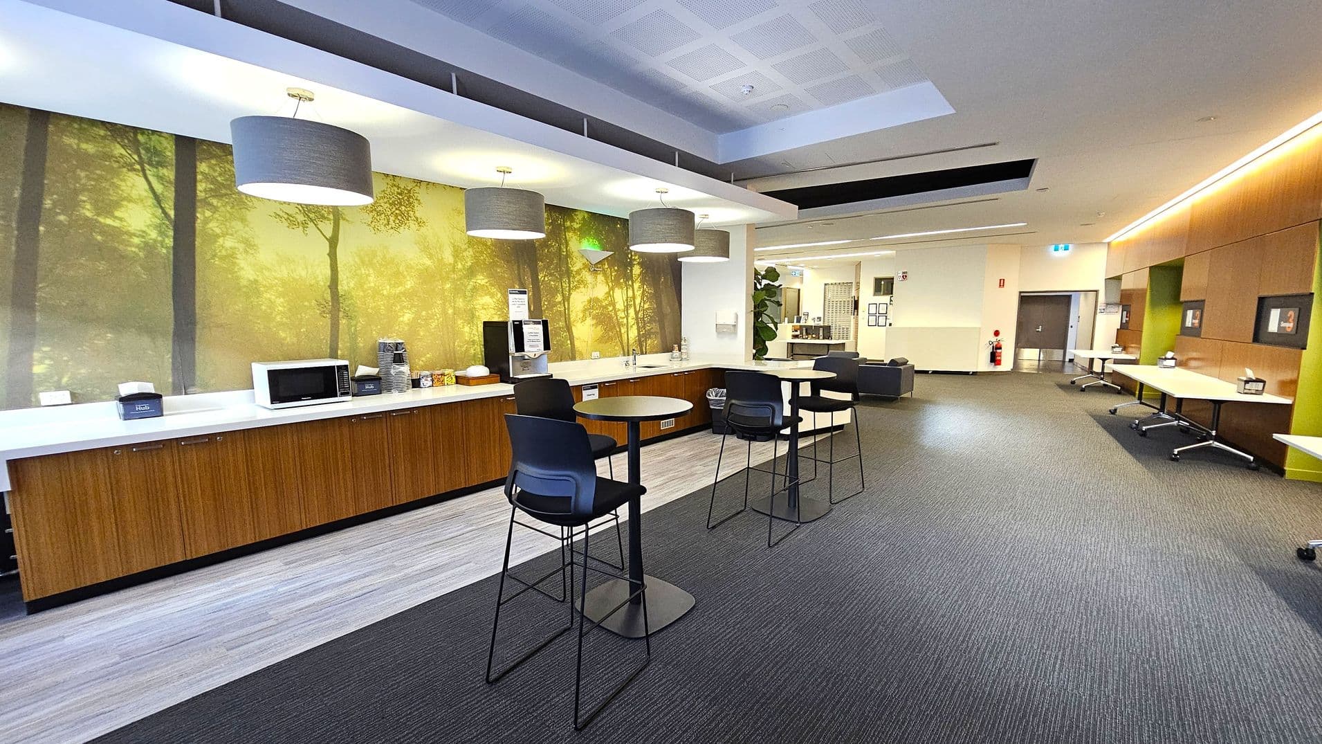 A modern break room with a forest-themed mural on the left wall. Below the mural, there is a long countertop with a microwave, coffee machine, and various supplies. Three high tables with black chairs are in the centre. The right side has a series of tables and a wall with wooden panelling. The ceiling features recessed lighting and large grey pendant lights. The floor is carpeted in a dark grey tone, and there is a fire extinguisher near the door in the background.