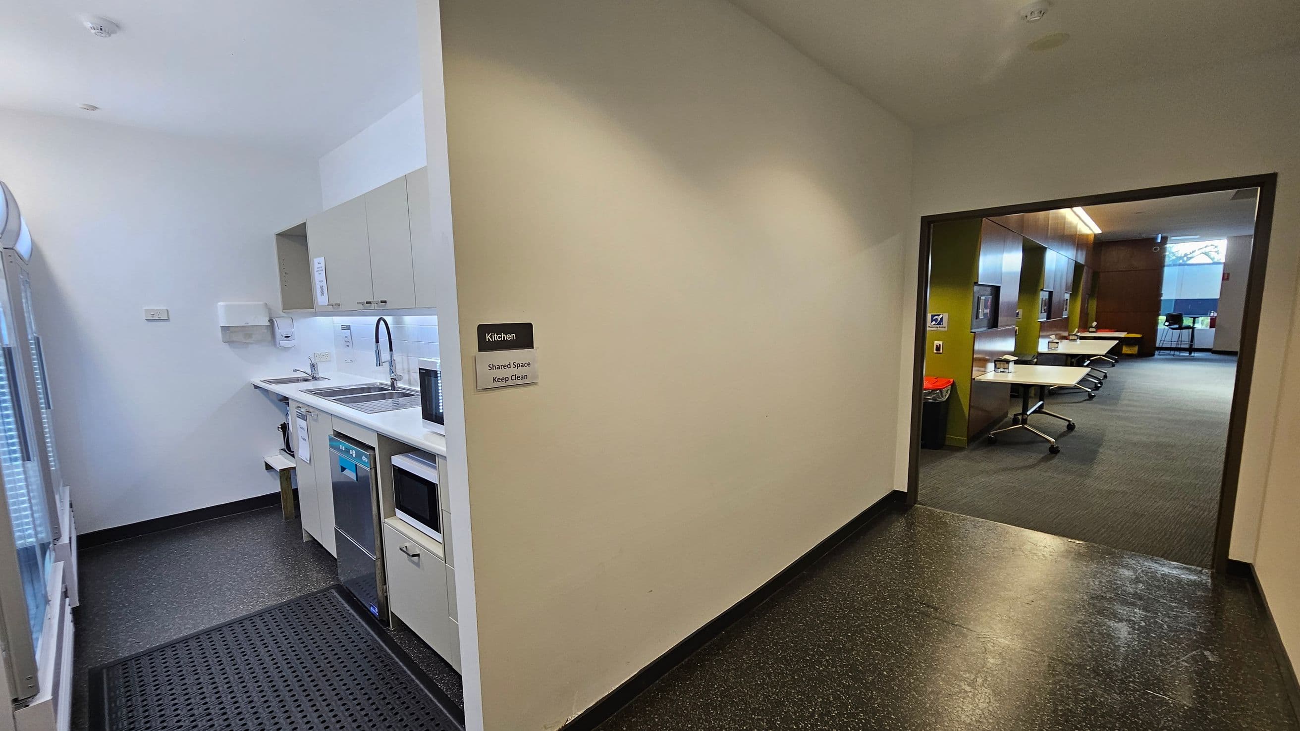 A small kitchen area is on the left with white cabinets, a sink, a microwave, and a dishwasher. A sign on the wall reads "Kitchen Shared Space Keep Clean." The floor is dark with a rubber mat near the entrance. On the right, an open doorway leads to a room with tables and chairs on a green carpet. The room has wooden panelling and large windows in the background.