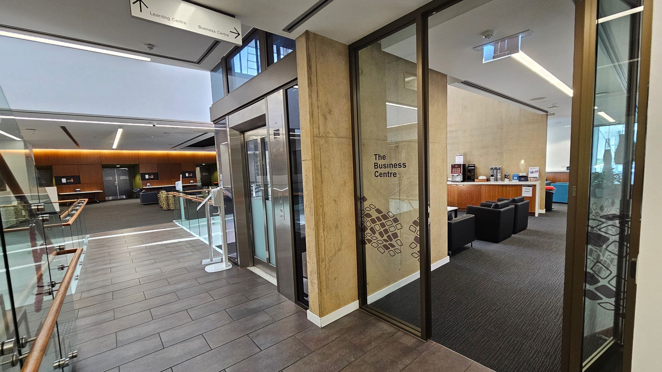 A modern interior with a corridor leading to a glass entrance labelled "The Business Centre" on the right. Inside, there are black chairs and a wooden counter with coffee machines. The floor is tiled, and the ceiling has recessed lighting. On the left, a glass railing lines the walkway, and a sign above points to the Learning Centre and Business Centre. The background features a spacious area with wooden panels and additional seating.