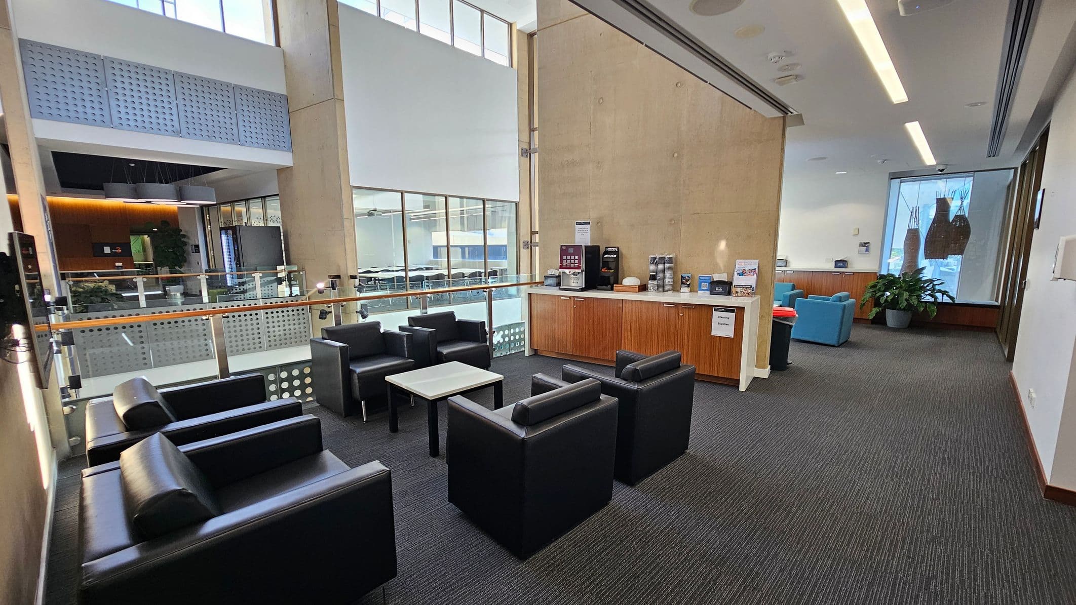 A modern lounge area with black leather chairs and a small table in the foreground. To the right, a wooden counter with a coffee machine and various supplies. The background features large windows allowing natural light, and a glass railing overlooking a lower level. On the far right, blue chairs and a potted plant are visible. The ceiling has recessed lighting, and the floor is carpeted in dark grey.
