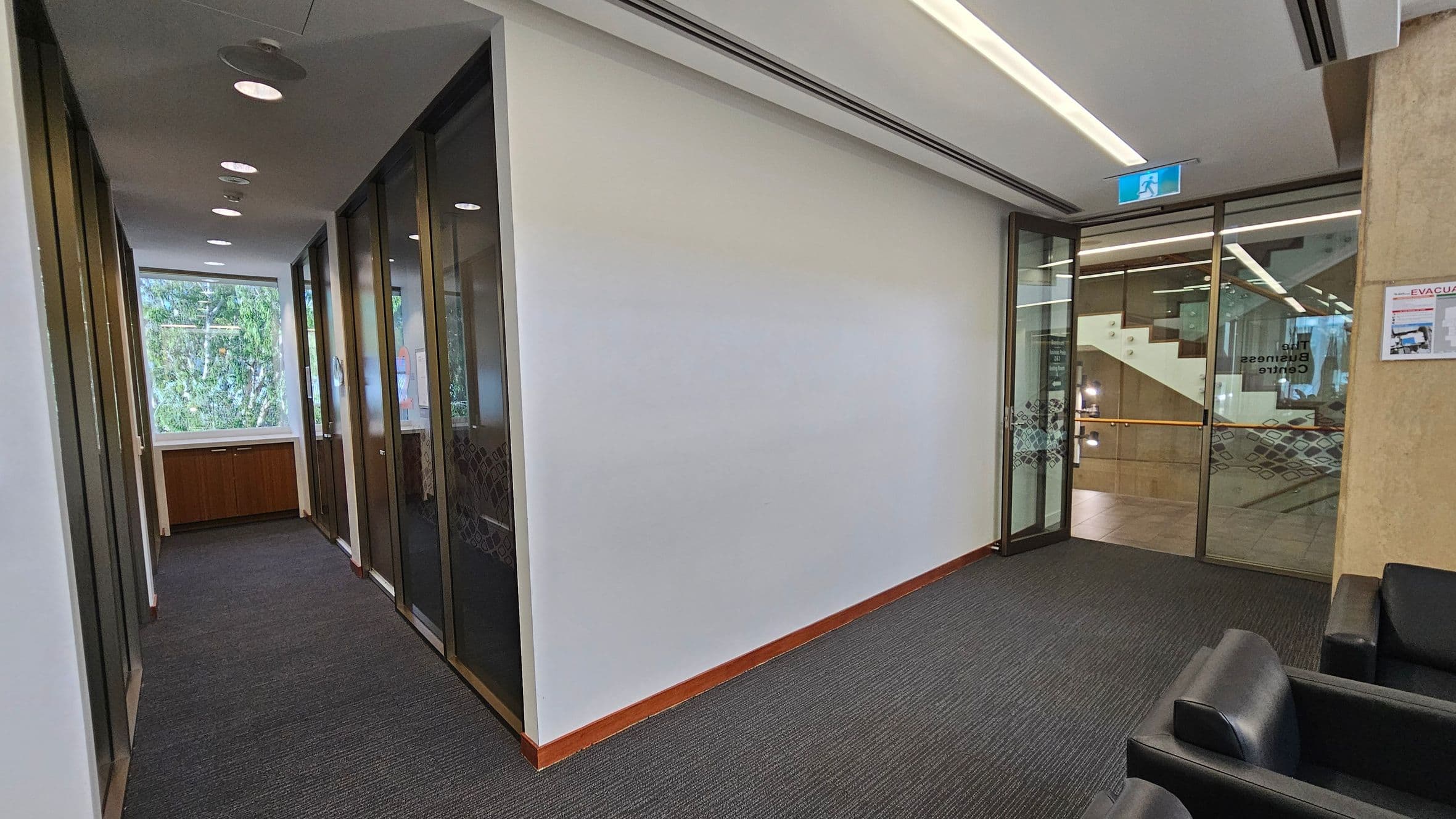A hallway with a dark carpeted floor and a white ceiling with recessed lighting. On the left, there are glass-panelled doors leading to offices, with a large window at the end showing greenery outside. On the right, a glass door with a sign reading "The Business Centre" leads to another area with visible stairs. A black leather chair is in the foreground, and an evacuation plan is mounted on the wall near the door.