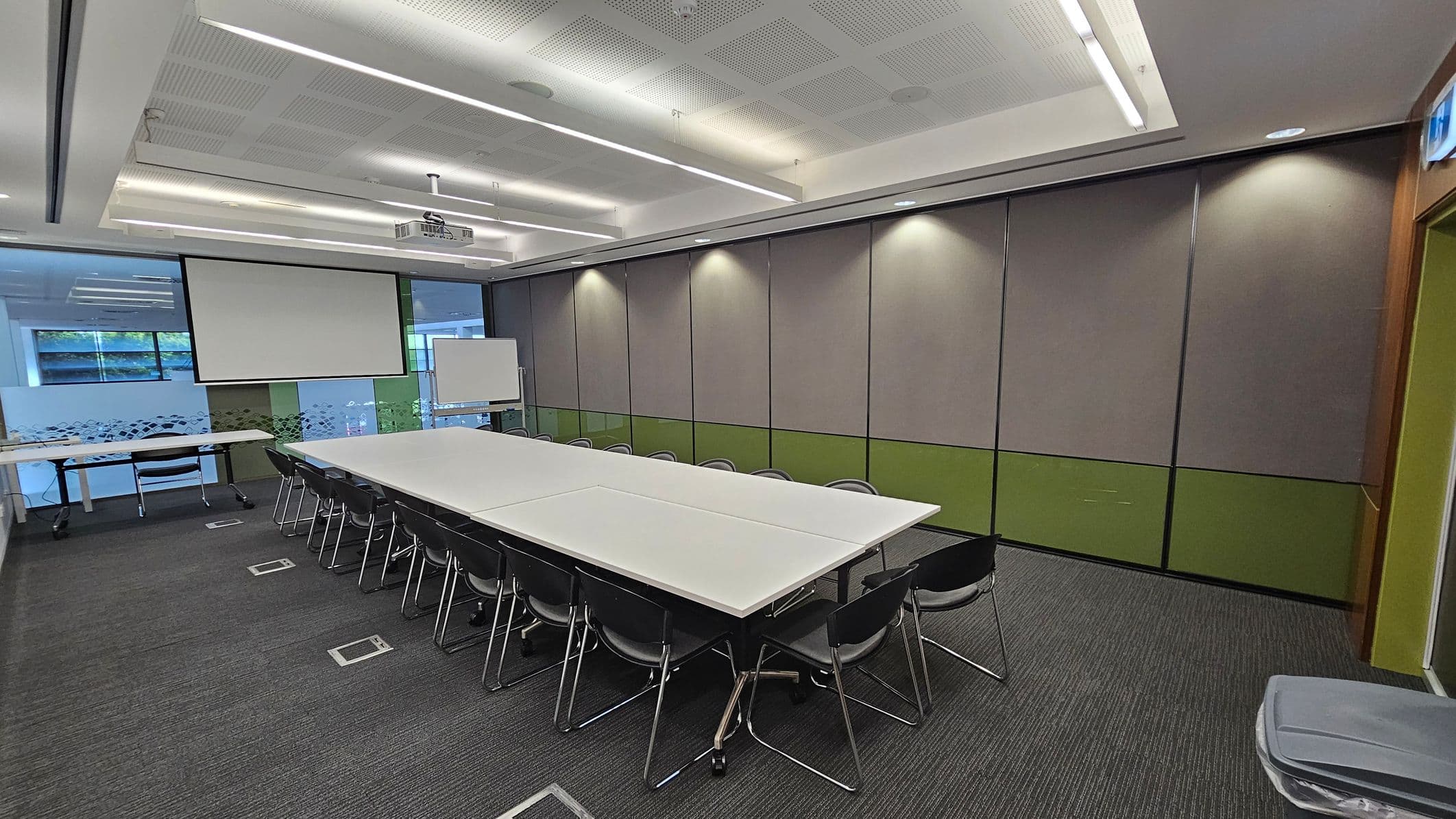 A conference room with a long rectangular table in the centre surrounded by black chairs. On the left, there is a large window with frosted glass and a pull-down projector screen. A whiteboard stands near the window. The walls are panelled with a mix of grey and green sections. The ceiling has recessed lighting and a mounted projector. The floor is carpeted in a dark grey tone.