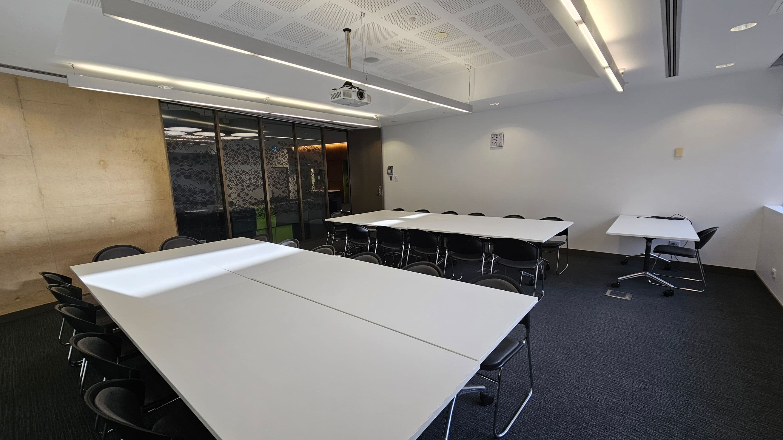 A modern conference room with white rectangular tables arranged in a U-shape, surrounded by black chairs. On the left, a wooden panelled wall is visible, while the right side features a plain white wall with a small clock. The back wall has glass panels with a patterned design. The ceiling is fitted with recessed lighting and a projector. The floor is carpeted in dark grey.