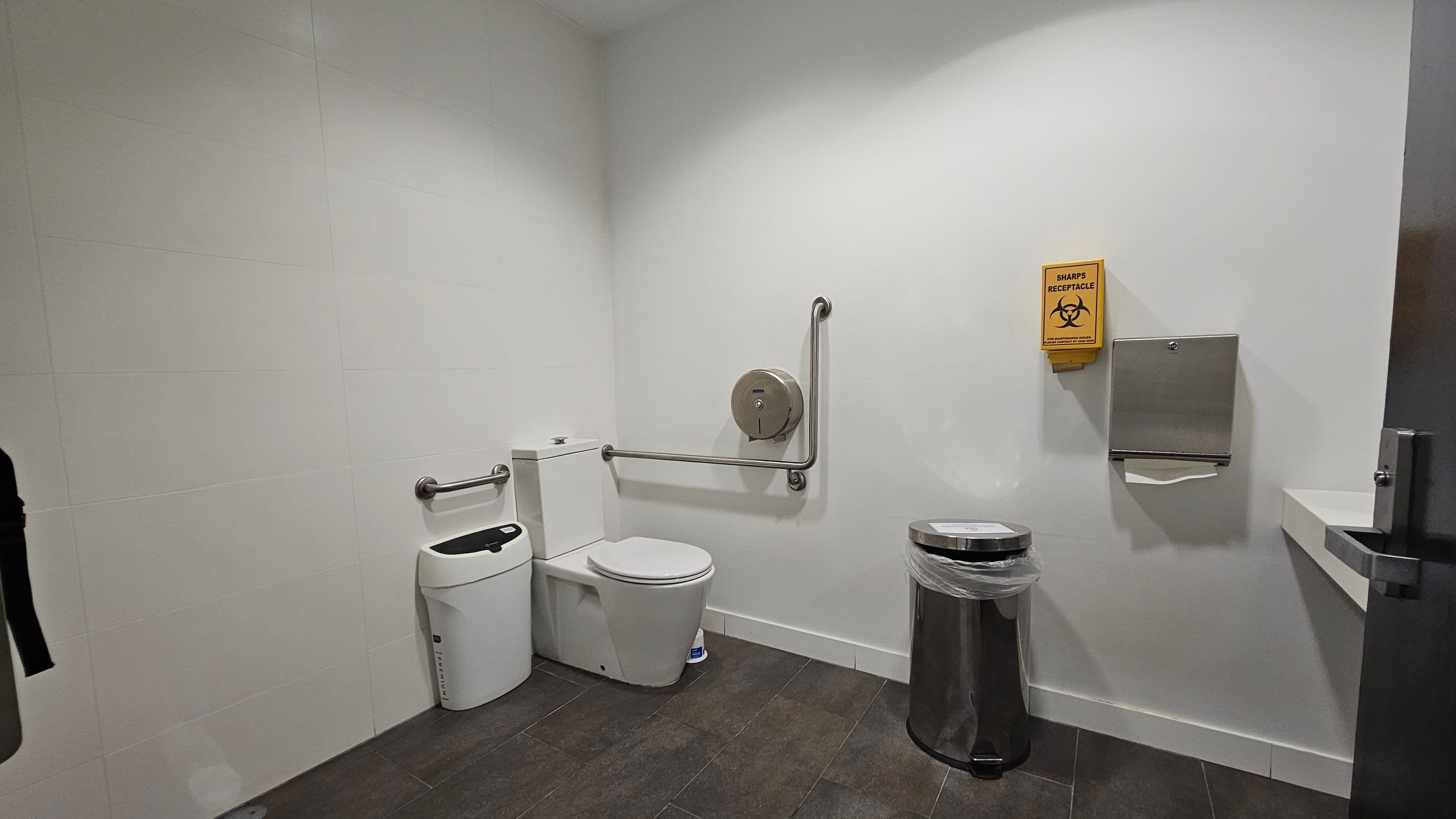 A public restroom with a white toilet and a sanitary bin on the left. Stainless steel grab bars are mounted on the walls near the toilet. A toilet paper dispenser is attached to the wall. On the right, there's a sharps receptacle with a biohazard symbol and a paper towel dispenser. A metal bin with a plastic liner is on the floor. The walls are white, and the floor is covered with dark tiles. A door with a metal handle is partially visible on the far right.