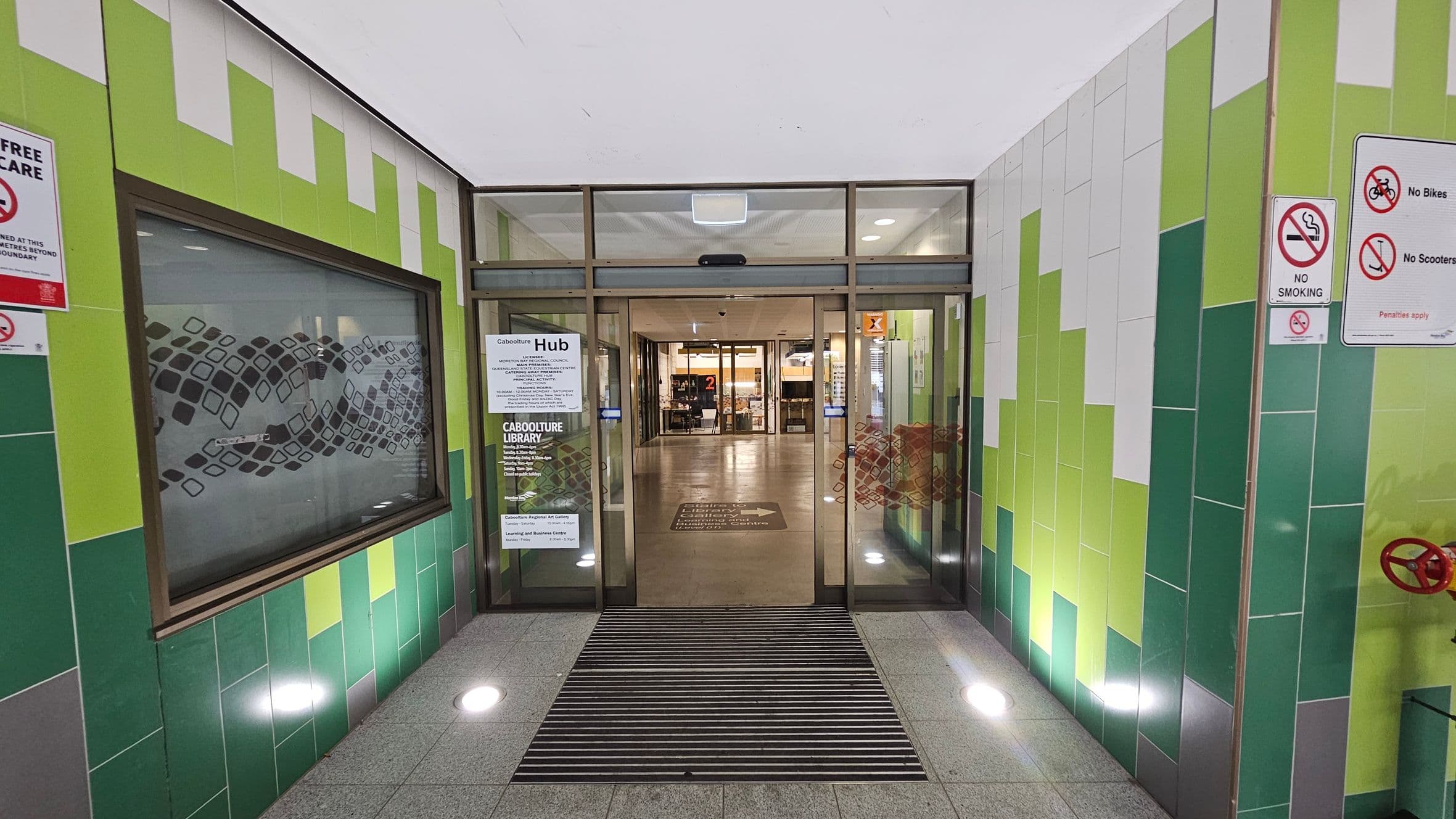 Entrance to a building with automatic glass doors in the centre. The walls are tiled in shades of green and white. On the left, a window with a decorative pattern. On the right, signs prohibiting smoking, bikes, and scooters. The floor is grey with a black mat in front of the doors. Inside, a well-lit hallway with visible signage and a number "2" in the distance. Ceiling lights are recessed and evenly spaced.