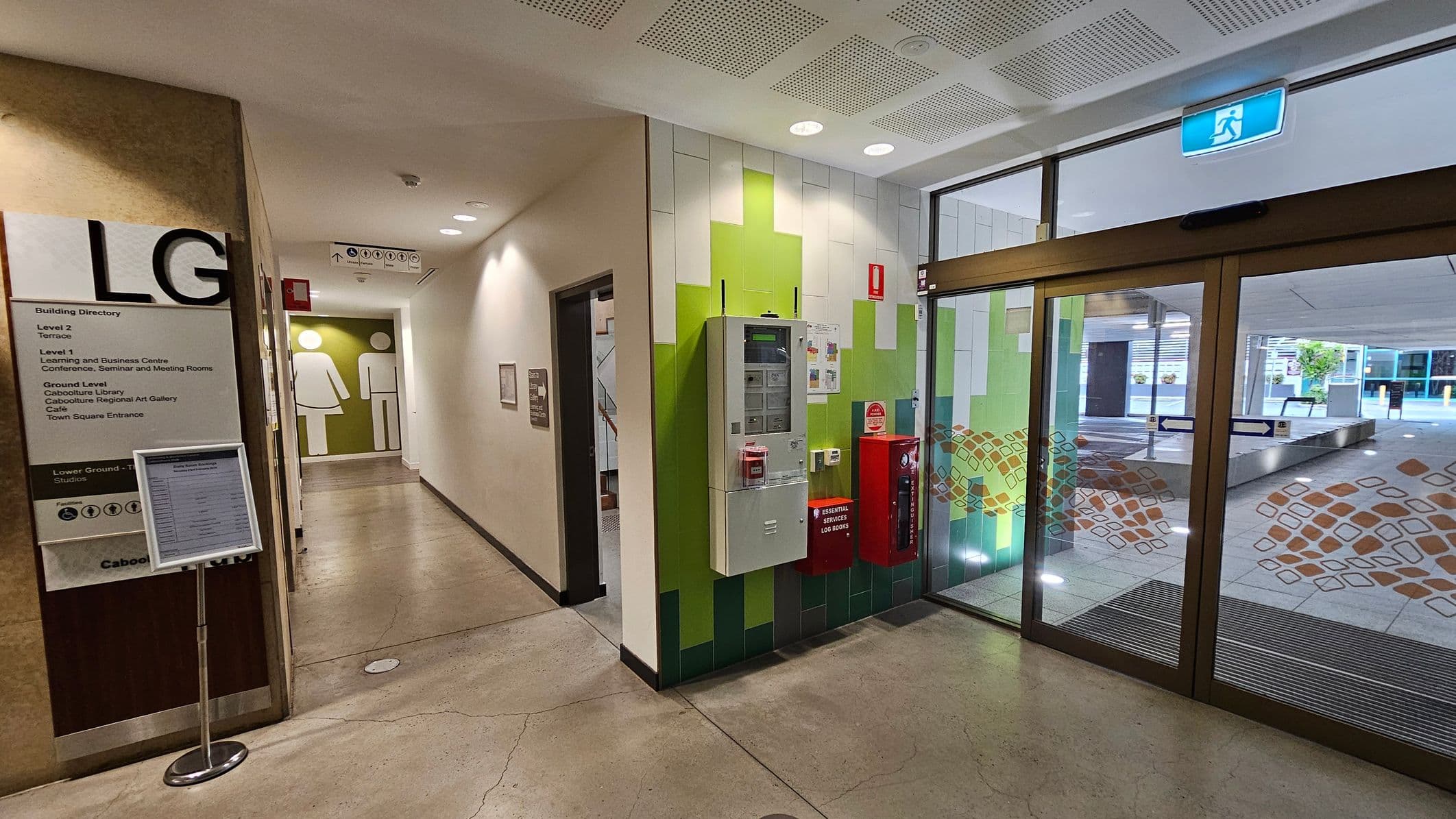 A hallway with a polished concrete floor. On the left, a building directory sign marked "LG" lists various levels and facilities. Further down, restroom signs are visible. On the right, a wall with green and white tiles features a control panel and fire safety equipment. Glass doors with decorative patterns lead outside, where a covered area and some greenery are visible. The ceiling has recessed lighting and ventilation panels.