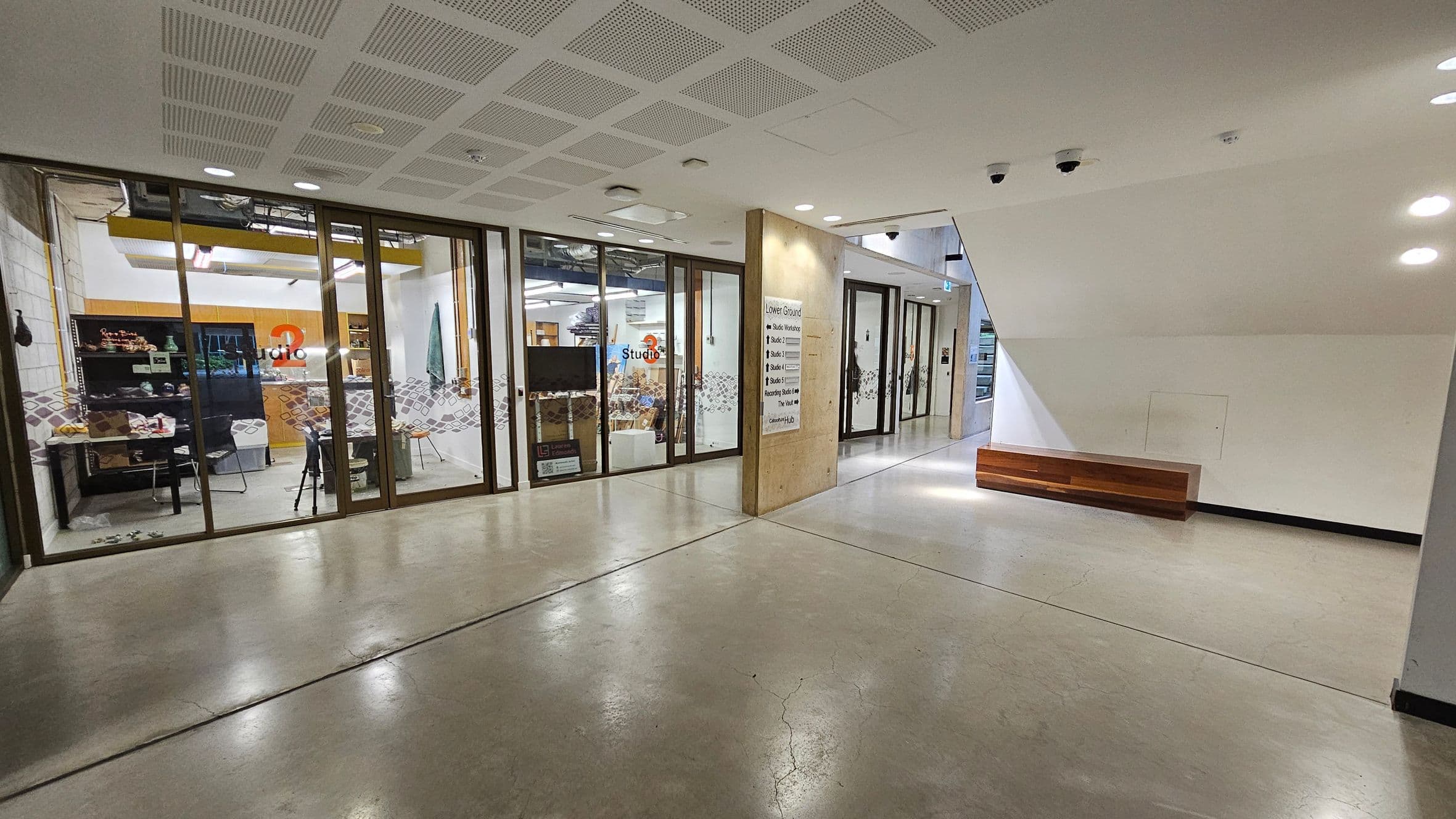 A modern interior space with a polished concrete floor and a ceiling with recessed lighting. On the left, there is a glass-walled studio with visible shelves and chairs inside. The studio has a sign with the number "3". In the centre, a concrete pillar displays directional signs. On the right, a white wall features a wooden bench attached to it. The corridor extends further back, with more glass doors and a continuation of the polished floor.