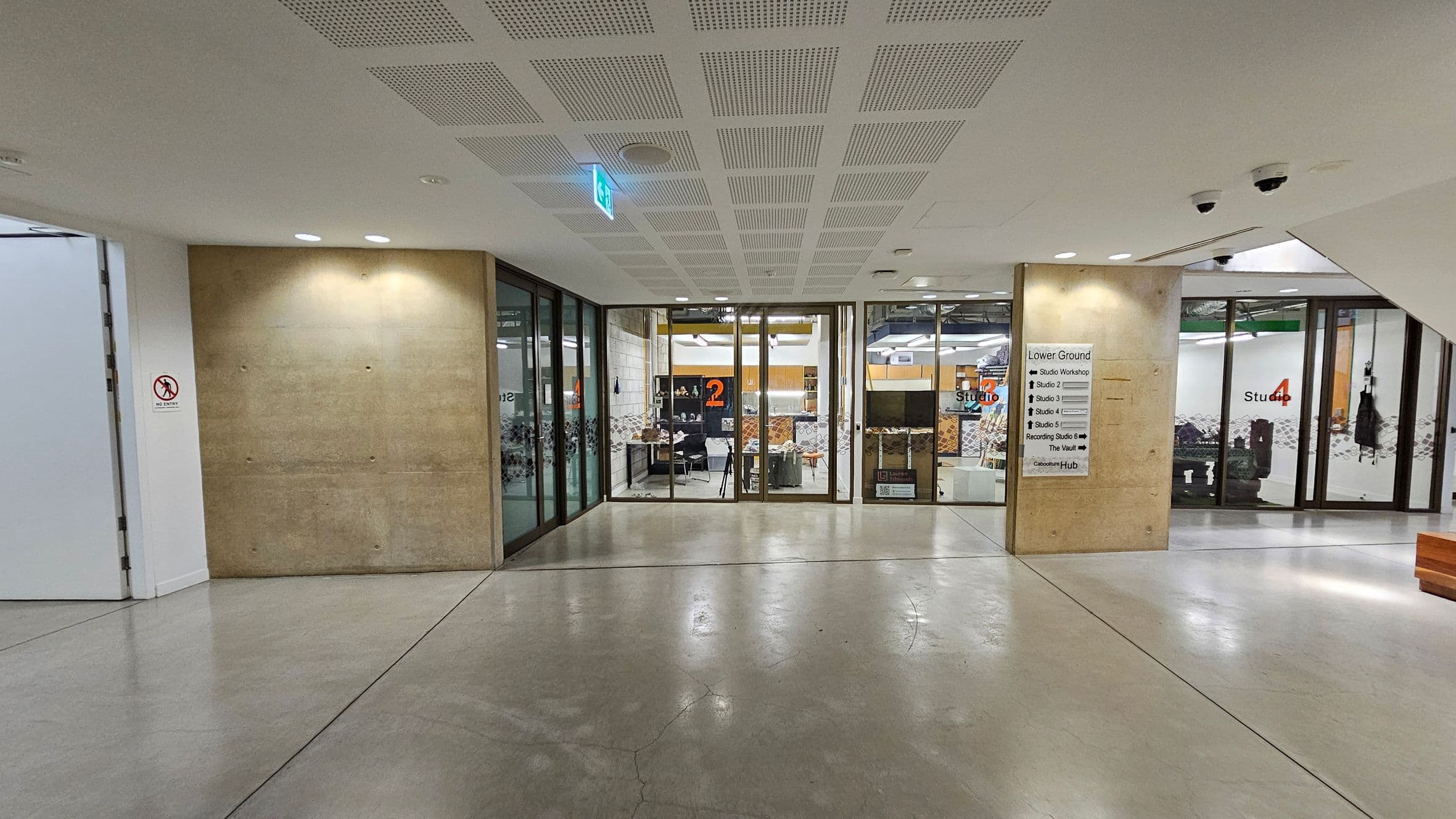 A hallway with a polished concrete floor and a perforated ceiling. On the left, a beige wall with a "no smoking" sign and glass doors leading to Studio 2. In the centre, a directory sign lists various studios and facilities. On the right, glass doors lead to Studio 4, with visible interior furnishings. Security cameras are mounted on the ceiling.