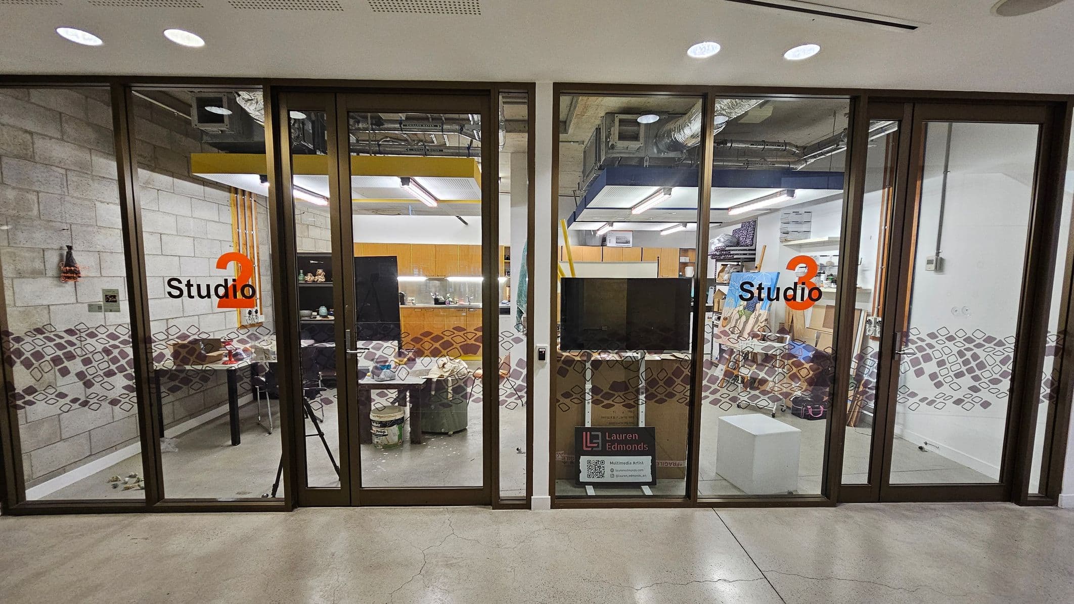 Glass-fronted studios labelled "Studio 2" and "Studio 3" with orange numbers. Inside, Studio 2 has a table with various items and a brick-patterned wall. Studio 3 contains art supplies, a canvas, and a white cube. A sign with "Lauren Edmonds" and QR codes is in front of Studio 3. The floor is polished concrete, and the ceiling has exposed ductwork and lighting fixtures.