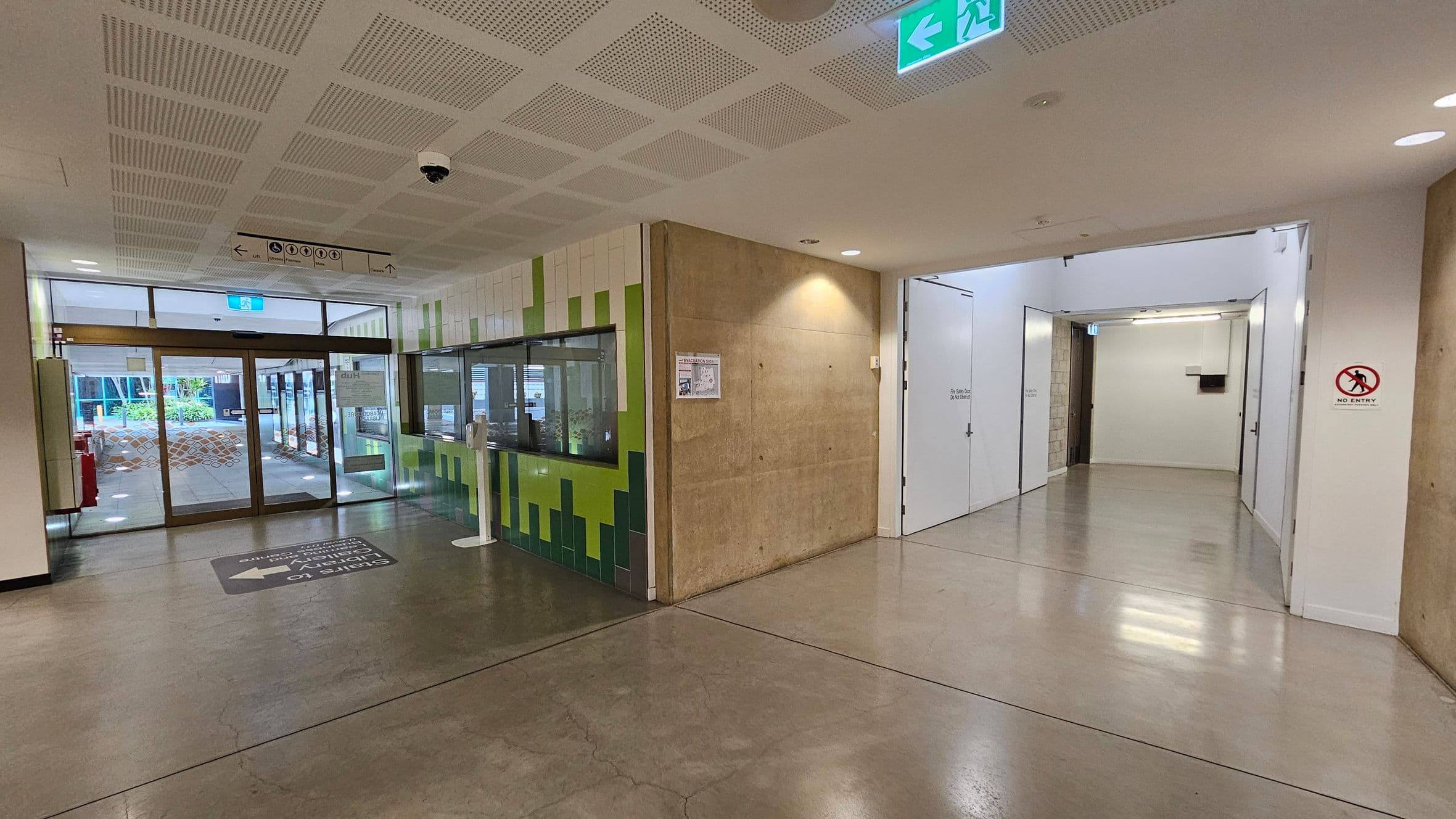 A hallway with a polished concrete floor and a perforated ceiling. On the left, there are glass doors leading outside, with a view of greenery. A wall with green and white tiles features a window and a sign. On the right, a corridor with white walls and a "No Entry" sign. An emergency exit sign is visible on the ceiling.