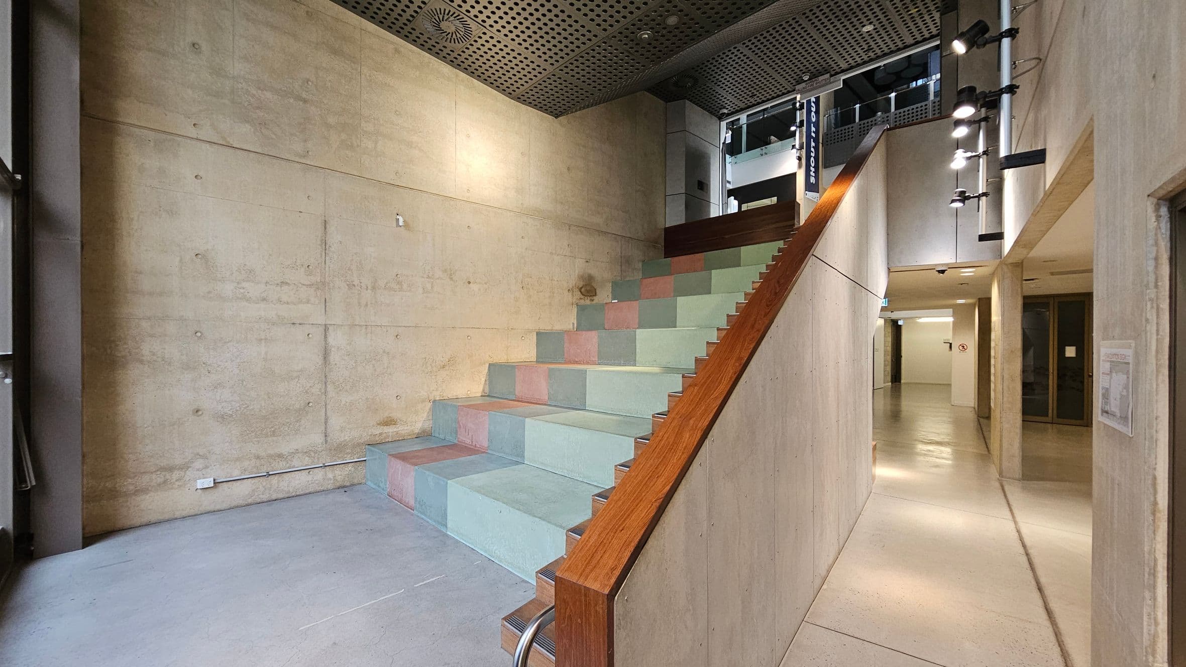 A modern interior space with a wide staircase in the centre, featuring multicoloured steps and a wooden handrail. The walls are concrete with visible textures and a few light fixtures on the right. The ceiling is perforated metal with recessed lighting. To the right, a corridor leads to another area, with doors and a no-smoking sign visible. The floor is smooth and light-coloured, complementing the industrial aesthetic.