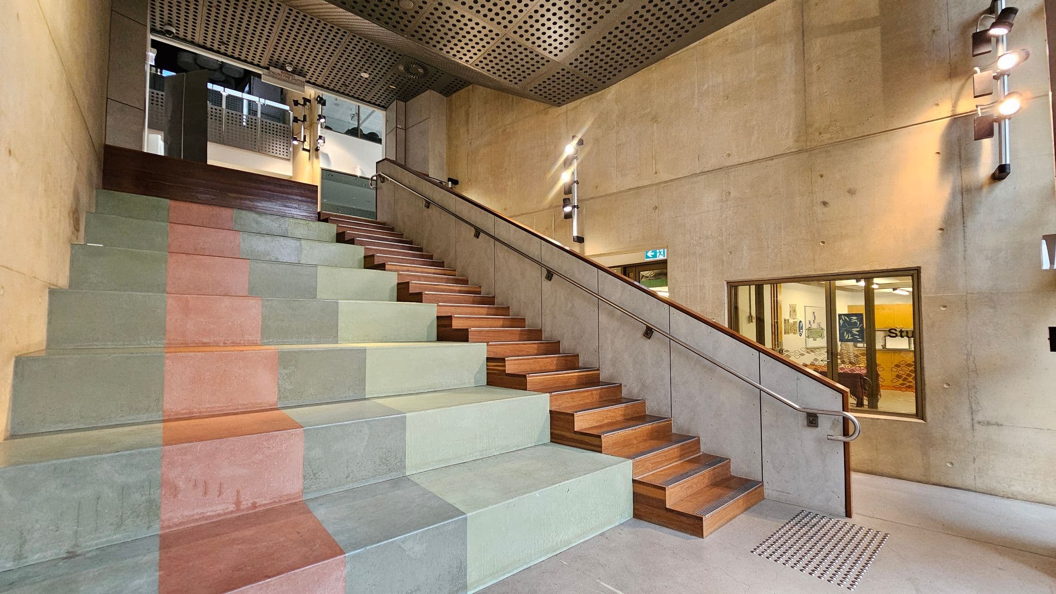 A modern interior with a wide staircase on the left featuring large, colourful steps in shades of green, orange, and grey. On the right, a narrower wooden staircase with a metal handrail runs parallel. The walls are concrete with mounted lights, and a window reveals a room with patterned flooring and wall art. The ceiling is perforated metal, and the floor is smooth concrete.
