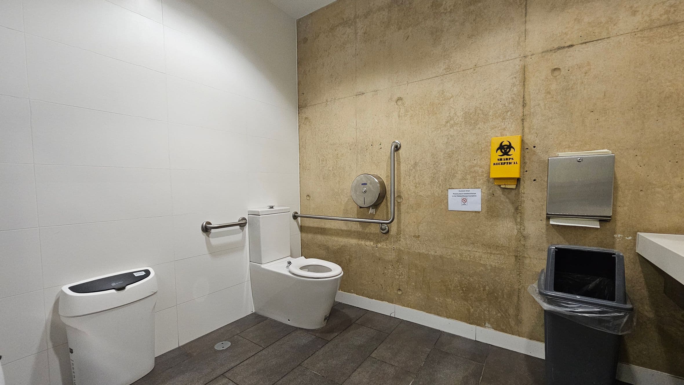 A public restroom with a white-tiled wall on the left and a brown textured wall on the right. In the centre, a white toilet with a metal grab bar beside it. Above the toilet, a large toilet paper dispenser is mounted. On the right wall, a yellow sharps disposal container and a paper towel dispenser are visible. Below them, a grey rubbish bin with a liner. The floor is covered with dark tiles.