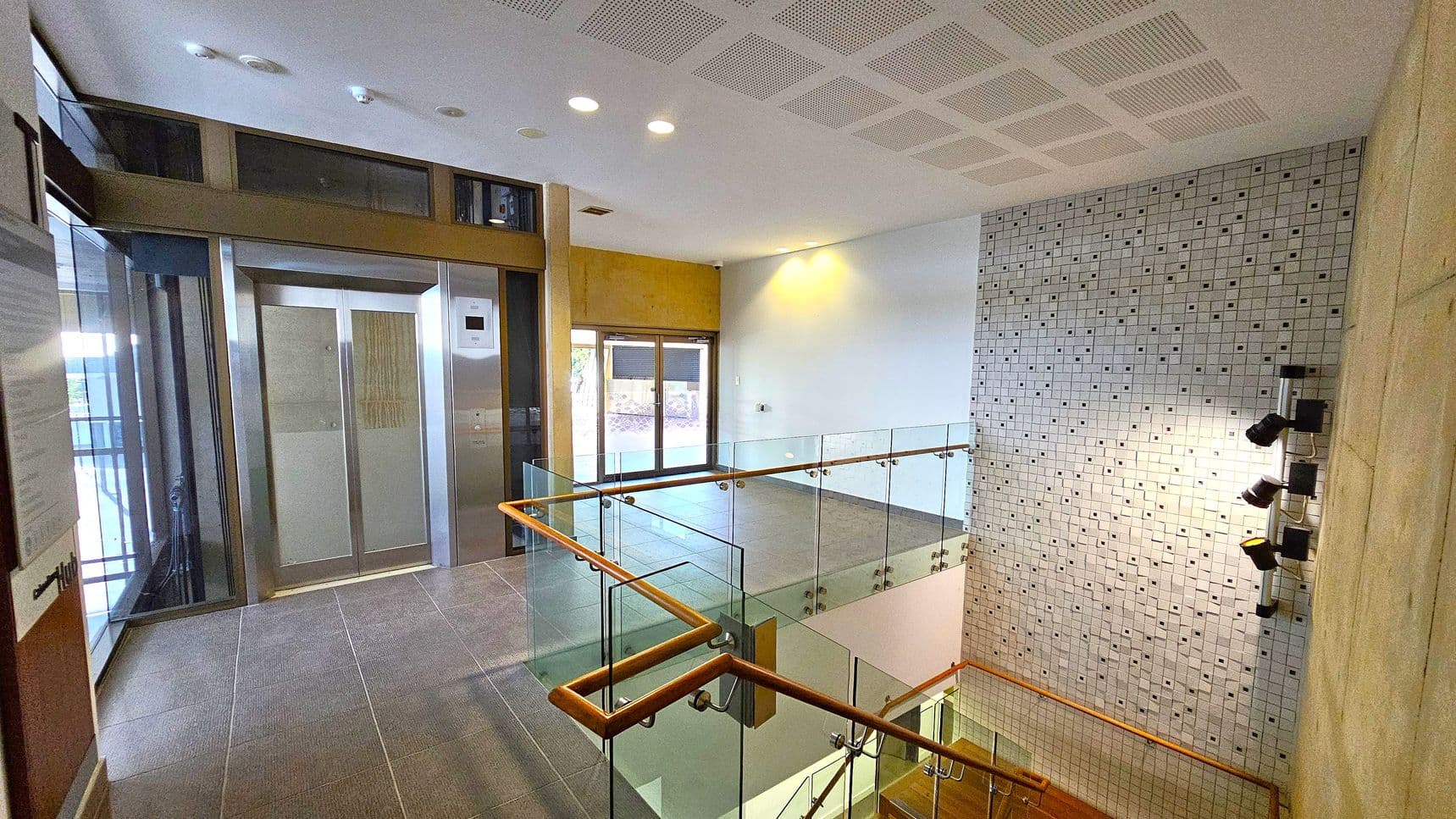 A modern indoor space with a lift on the left, featuring metallic doors and a control panel. In the centre, a glass railing with wooden handrails surrounds a staircase leading downwards. The right wall is covered with a mosaic of small square tiles in various colours. The ceiling is white with recessed lighting, and the floor is tiled in grey. A set of spotlights is mounted on the wall near the staircase.