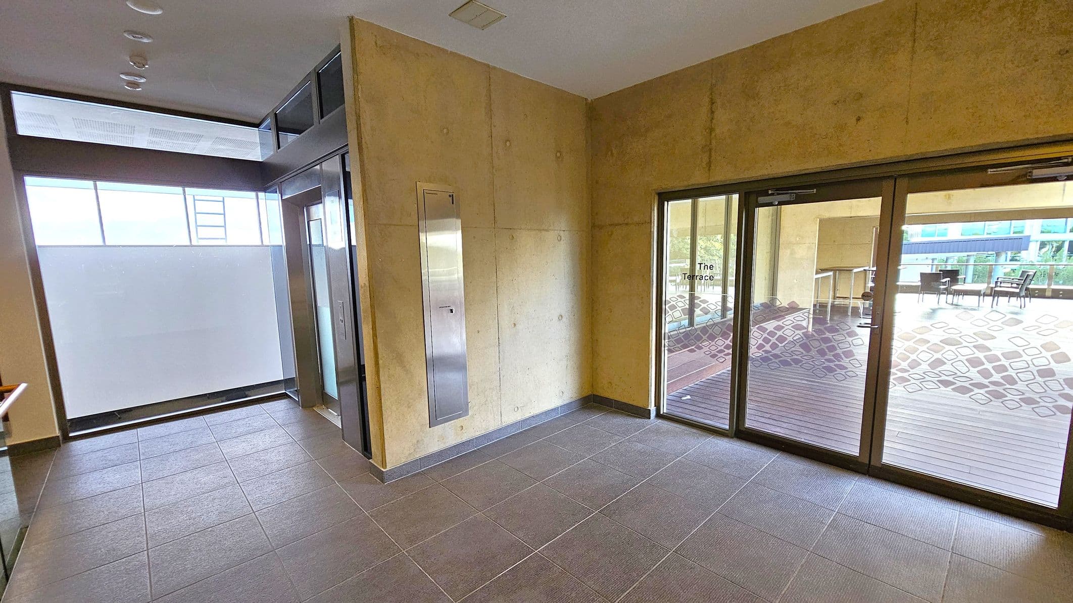 A modern indoor space with grey tiled flooring. On the left, there's a lift with a metallic door and a frosted glass window above it. The right side features large glass doors leading to an outdoor terrace area, with chairs and tables visible. The walls are a light beige colour, and the ceiling is plain with recessed lighting. The terrace has a wooden deck and decorative patterns on the glass doors.