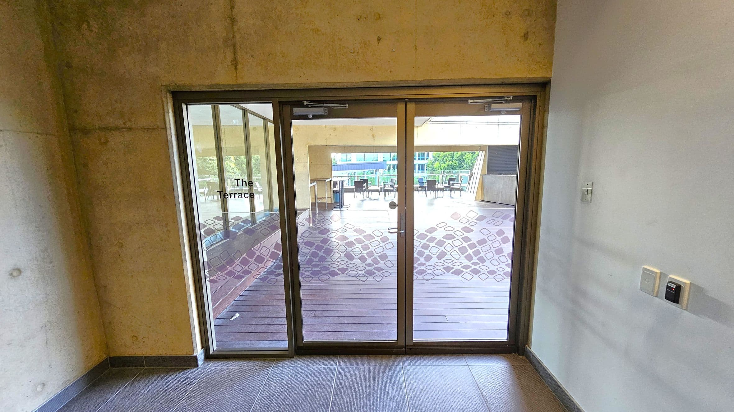 Glass doors with a geometric pattern lead to an outdoor terrace. The left wall is concrete, while the right wall is painted white with a light switch and control panel. The floor inside is tiled, transitioning to wooden decking outside. The terrace has tables and chairs, with a view of greenery and a building in the background.