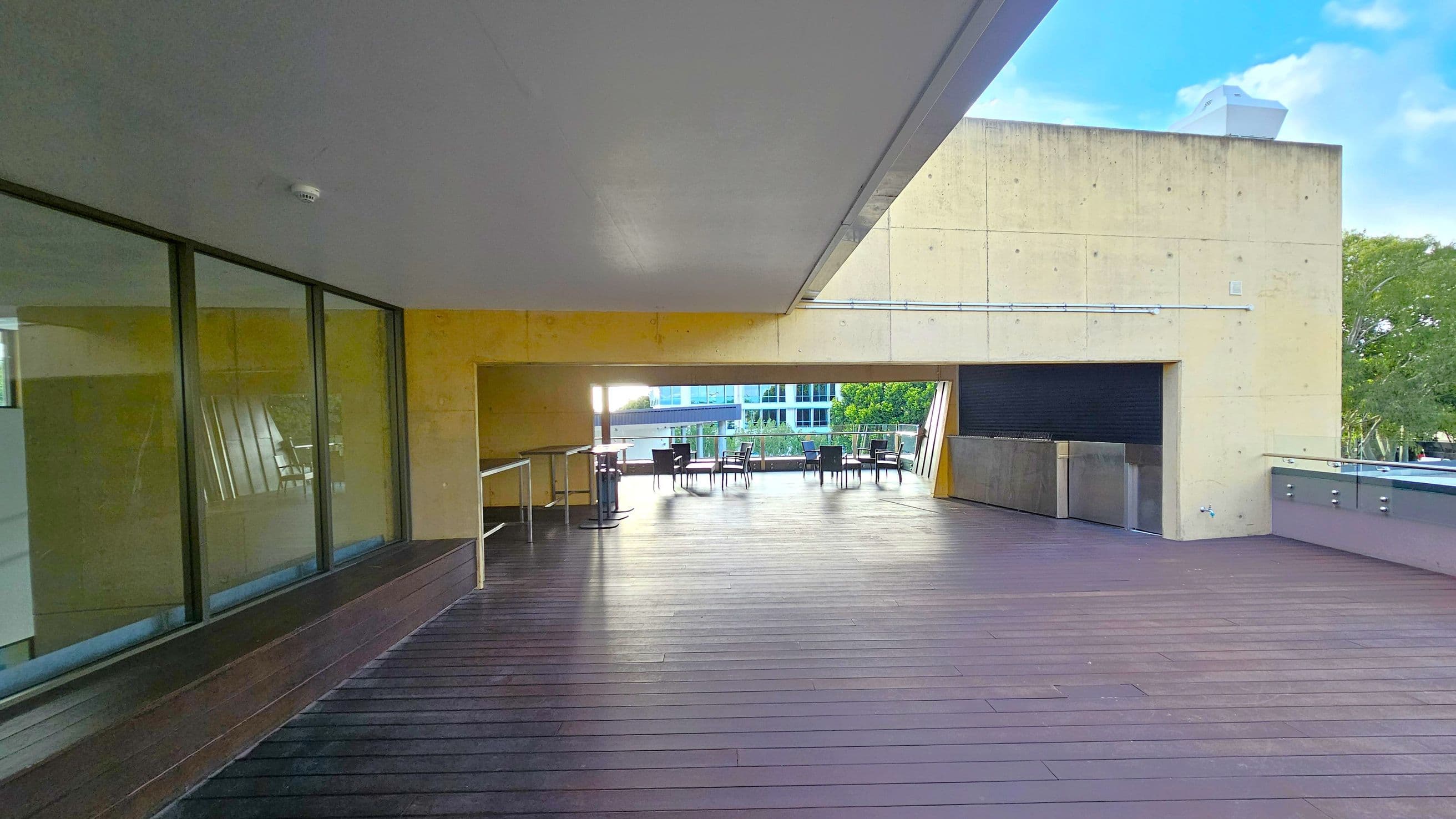 A spacious outdoor terrace with a wooden floor and a concrete ceiling. On the left, there are large glass windows reflecting the surroundings. In the centre, several tables and chairs are arranged near a railing, offering a view of trees and a building in the background. On the right, a concrete wall with a closed shutter and a countertop is visible. The area is partially covered, with open space allowing natural light to illuminate the terrace.