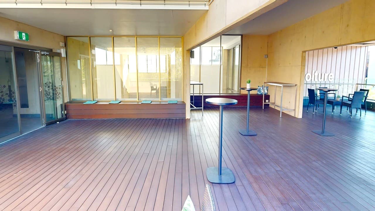 A spacious indoor area with wooden flooring. On the left, there are glass doors and a wooden bench with cushions. In the centre, tall round tables are placed on the floor. On the right, there's a bar counter with a bottle and a small plant. Further right, there are tables and chairs arranged in a seating area. The walls are light-coloured, and the ceiling is plain. Large windows in the background allow natural light to enter.