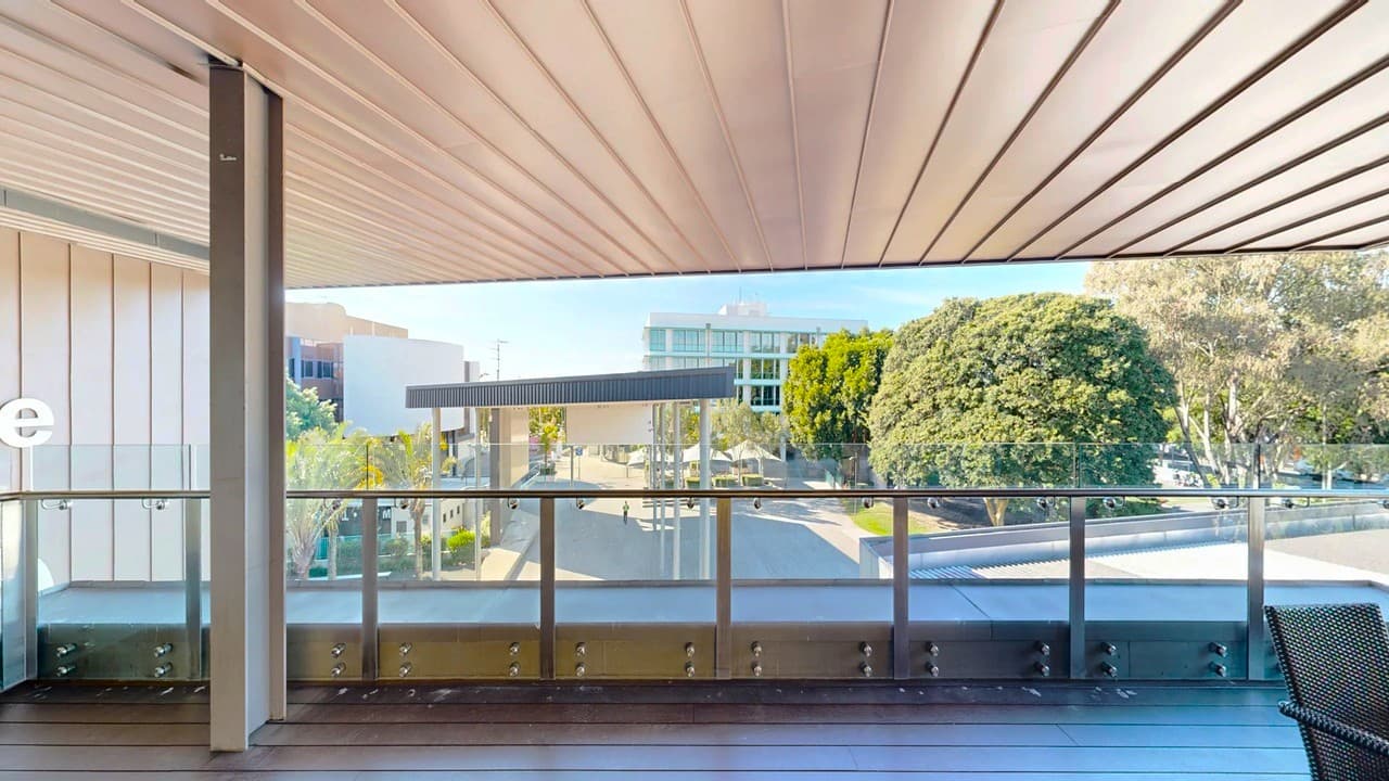 A modern balcony with a wooden floor and a glass railing. The ceiling is lined with parallel wooden panels. In the background, there are trees and a few buildings, including a multi-storey structure. A shaded area with a flat roof is visible, and there are people walking below. The scene is bright and open, suggesting a sunny day.