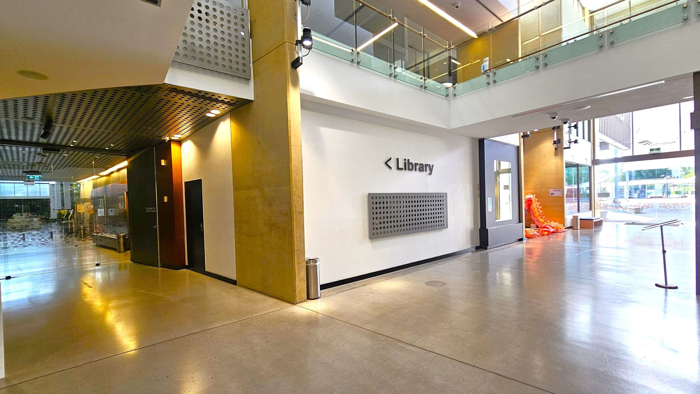 A modern interior space with polished concrete floors and a perforated metal ceiling. On the left, there's a corridor with glass walls and a dark door. In the centre, a white wall displays a sign pointing to the library. On the right, large windows allow natural light in, and an orange sculpture is visible near the entrance. Above, a glass railing lines the upper level.