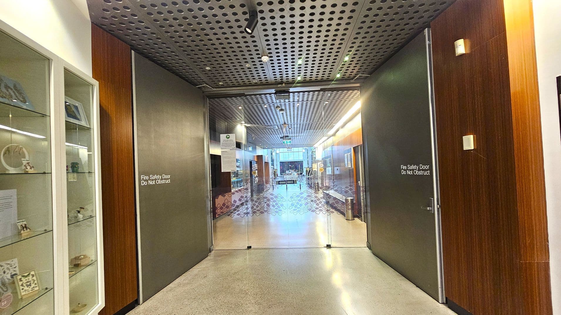 A corridor with a polished concrete floor and a perforated metal ceiling. On the left, there is a glass display cabinet with various items. In the centre, a glass door with a patterned design leads to a well-lit hallway. On the right, a dark fire safety door with a "Do Not Obstruct" sign is set in a wooden panel. The hallway beyond is lined with more wooden panels and display cases, with a visible exit sign in the distance.