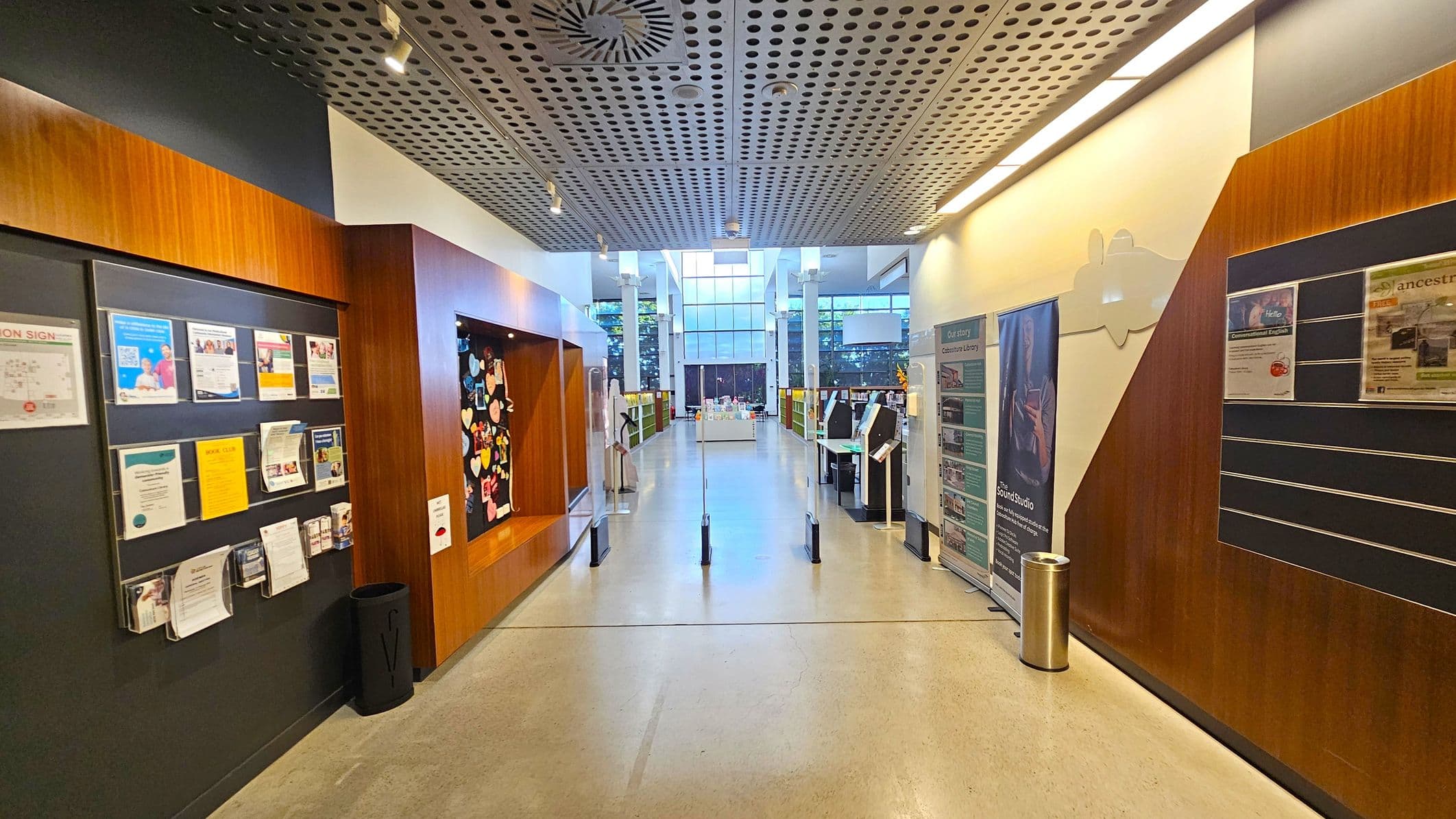 A hallway with a polished floor and a perforated ceiling. On the left, there are noticeboards with various flyers and a black bin. On the right, more noticeboards and a cylindrical metal bin are visible. The walls are adorned with wooden panels. In the centre, a pathway leads to a well-lit area with large windows and bookshelves. There are informational stands and a display table in the background.