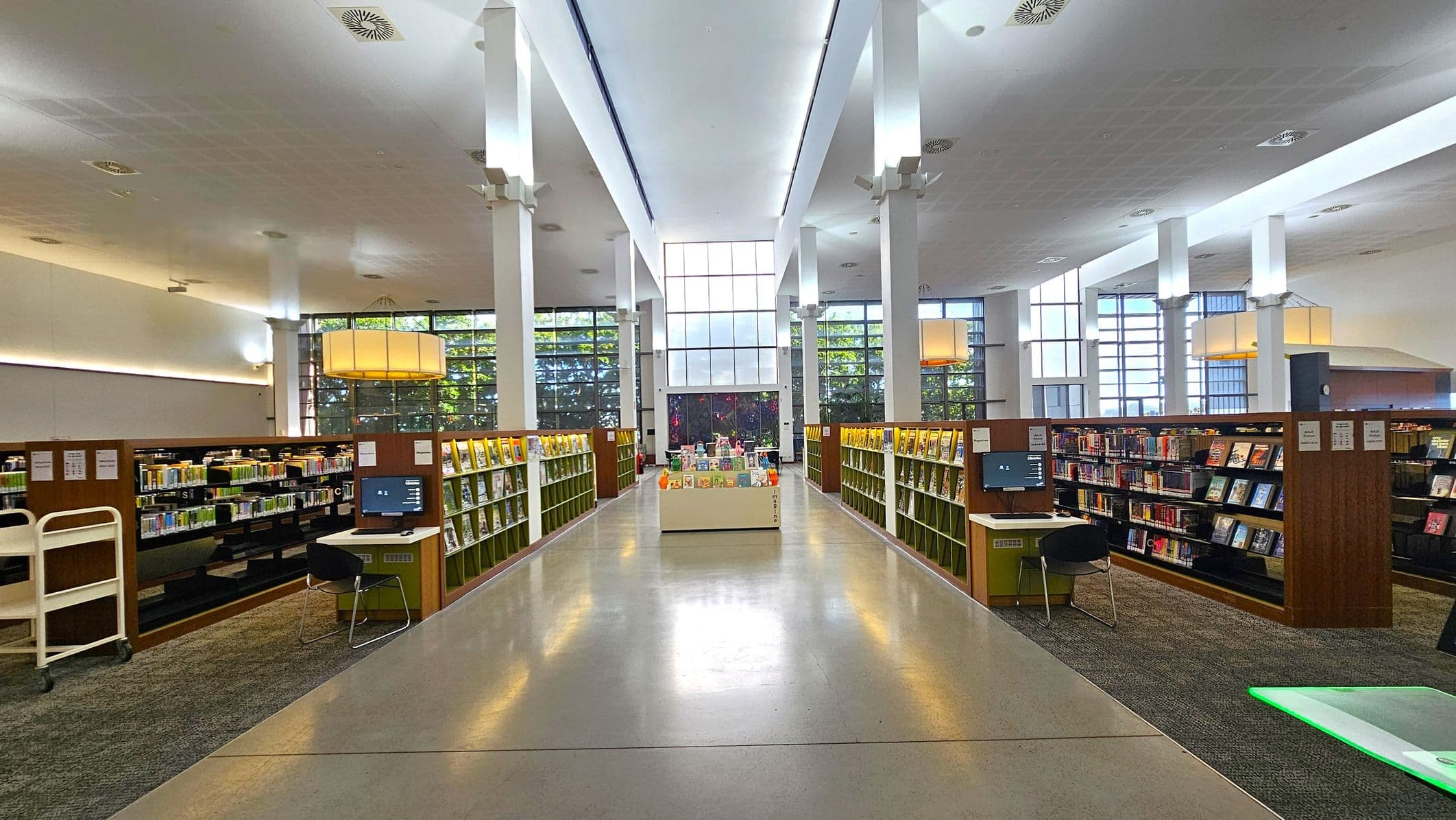 A spacious library interior with tall white columns and a high ceiling. In the centre, a display table holds various books. On both sides, there are rows of bookshelves filled with books. Each side has a computer station with a chair. Large windows in the background allow natural light to illuminate the space. The floor is a polished light grey, and the ceiling features recessed lighting.