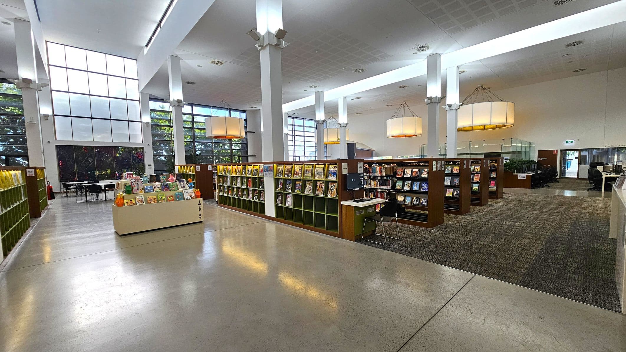A spacious library interior with high ceilings and large windows on the left, allowing natural light to flood in. In the centre, there are rows of bookshelves filled with books and magazines. A display stand with children's books is in the foreground. On the right, there are more bookshelves and a seating area with computers. The floor is a mix of polished concrete and carpet, and large pendant lights hang from the ceiling.