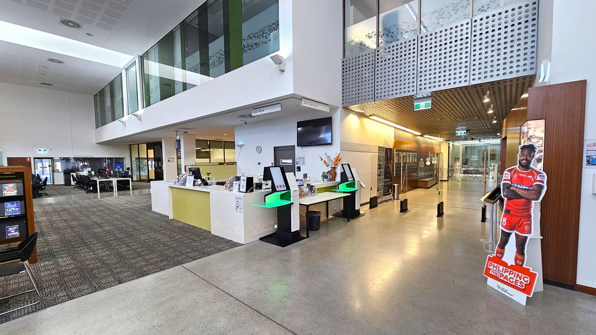 A spacious interior with a high ceiling and a mix of carpeted and polished concrete flooring. On the left, there is a seating area with tables and chairs. In the centre, a reception desk with computers and a "Self Service" sign above. To the right, a life-sized cardboard cutout of a person in a red sports uniform stands near a wooden panelled wall. The background features glass walls and a corridor with display cases. The ceiling has recessed lighting and a modern design.
