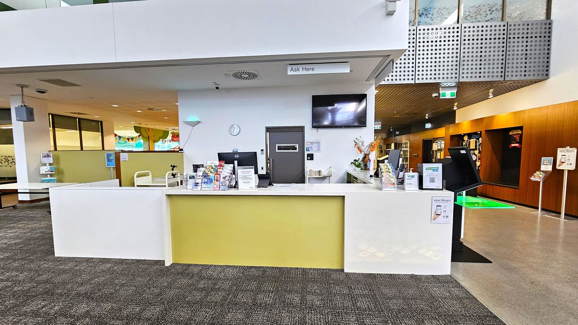 A modern library reception area with a white and yellow counter in the centre. On the counter are brochures, a computer, and a sign that reads "Ask Here". Behind the counter, a wall-mounted clock and a television screen are visible. To the left, there are partitions and a colourful mural with tree designs. On the right, a wooden wall with display shelves and a green-lit floor section. The floor is carpeted, and the ceiling has a mix of white and perforated metal panels.