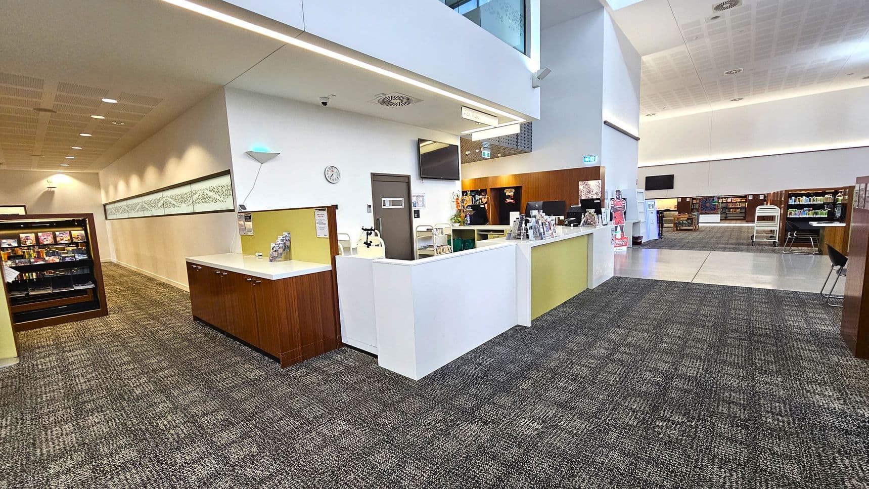 A spacious interior with a modern design. On the left, there is a wooden display case with shelves containing various items. The middle features a reception desk with white countertops and brochures. A clock and a wall-mounted TV are above the desk. The right side opens into a larger area with bookshelves and seating. The floor is carpeted with a patterned design, and the ceiling is high with recessed lighting.