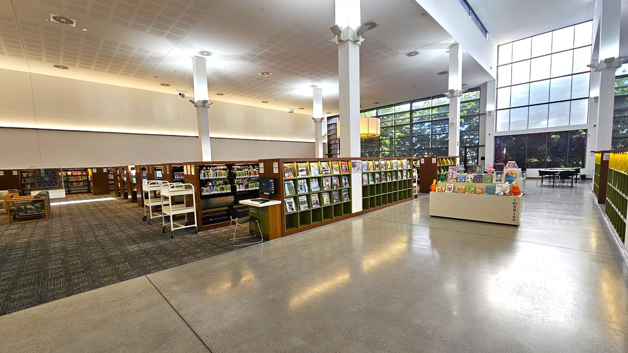 A spacious library interior with a polished concrete floor. On the left, there are rows of bookshelves filled with books, and a few seating areas with armchairs. A book cart is visible near the shelves. In the centre, a display stand showcases children's books. On the right, more bookshelves line the wall, and a few tables and chairs are set up near large windows. The ceiling is high with white pillars and recessed lighting.