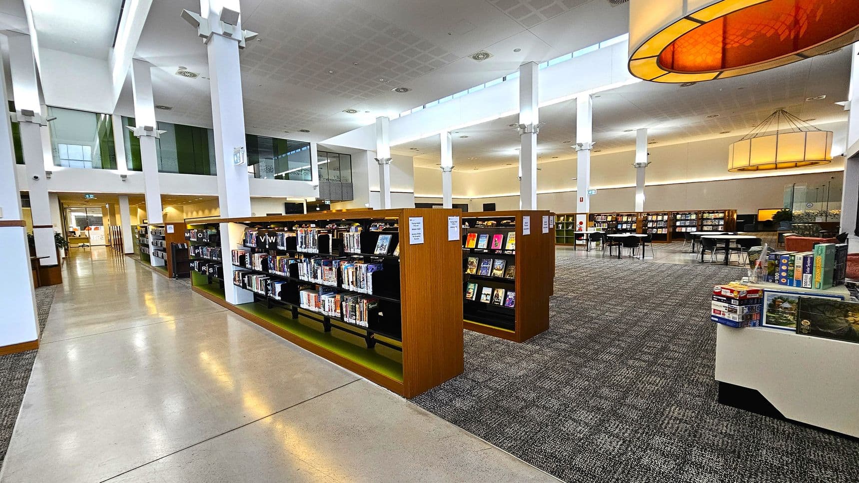 A spacious library interior with tall white columns and a high ceiling. On the left, there are bookshelves filled with books, and a polished concrete floor. In the centre, more bookshelves are arranged neatly, with a carpeted area. On the right, there are tables and chairs for reading, and a display with board games. Large, circular light fixtures hang from the ceiling, providing illumination. The background features more bookshelves and a glass-walled area.